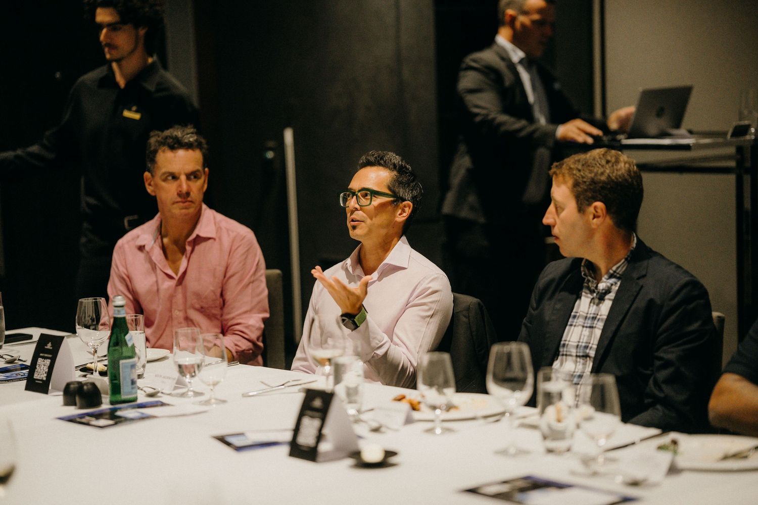 Three professionals engaged in discussion at a formal dinner table with wine glasses and place settings.