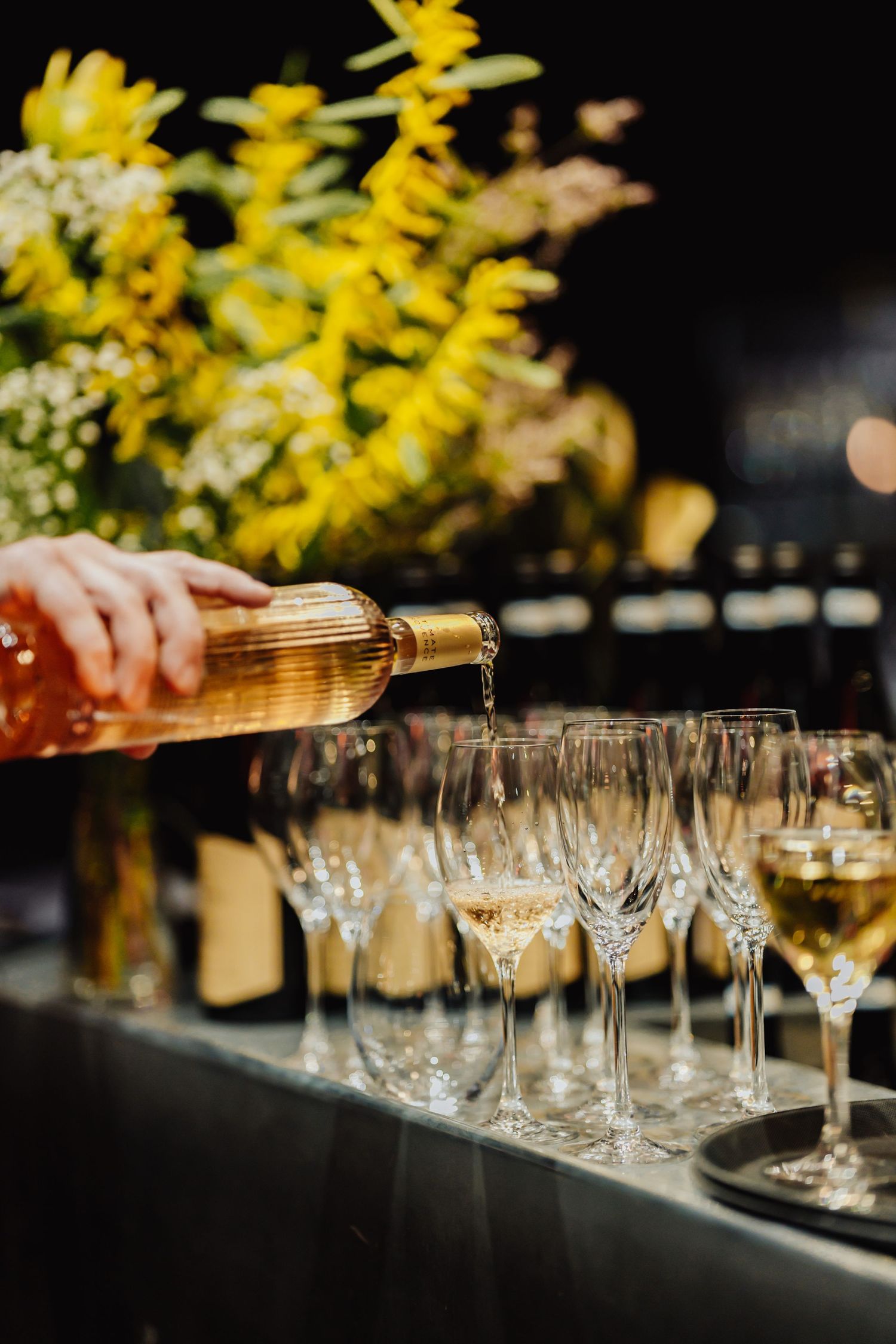 A row of empty wine glasses being filled with golden liquid from a bottle against a dark background with yellow flowers.