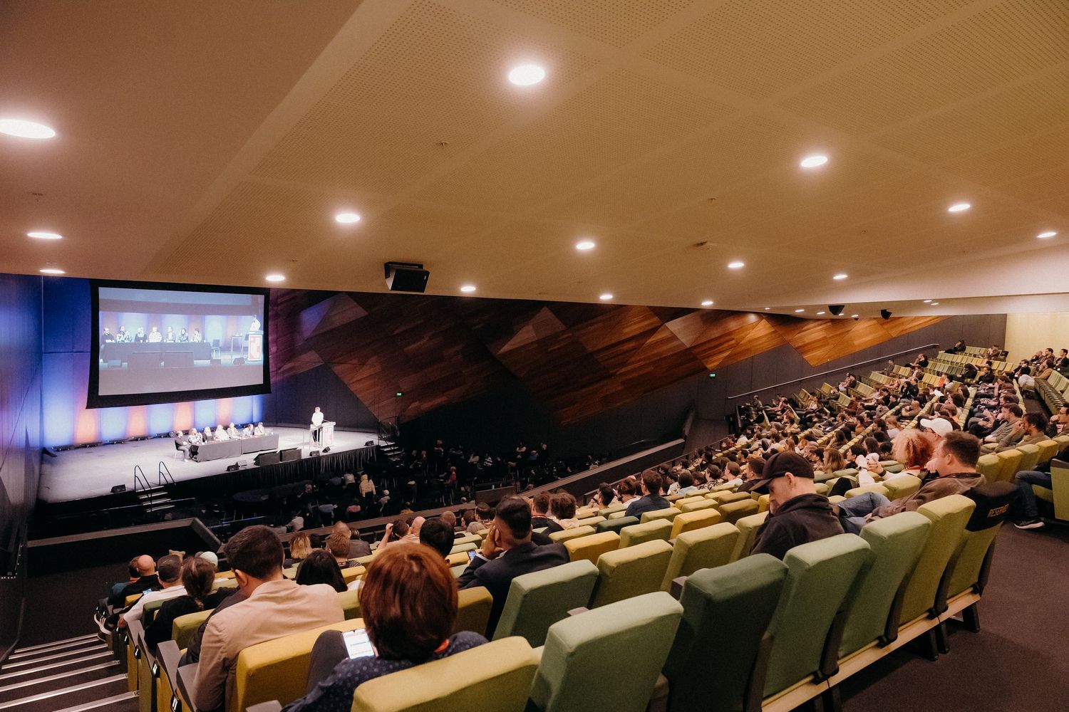 Large audience seated in a sloped auditorium with stadium seating facing a presentation screen and stage area.