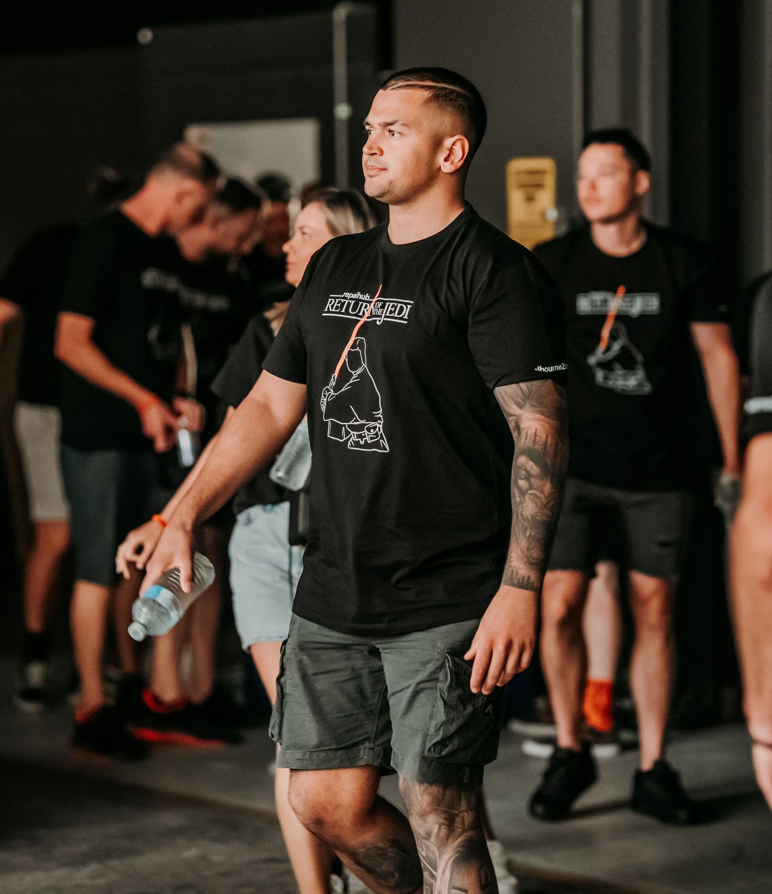 Athletic person in black t-shirt and cargo shorts holding water bottle stands among others in indoor workout setting.