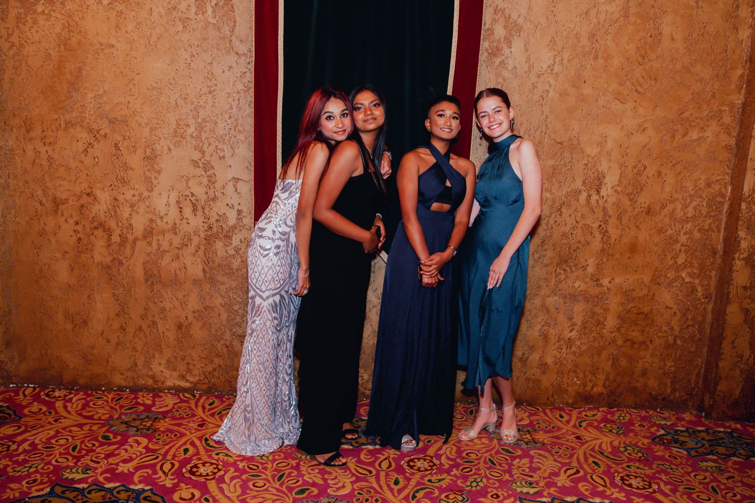 Four women in elegant evening gowns pose together against a textured gold wall on a red carpet.