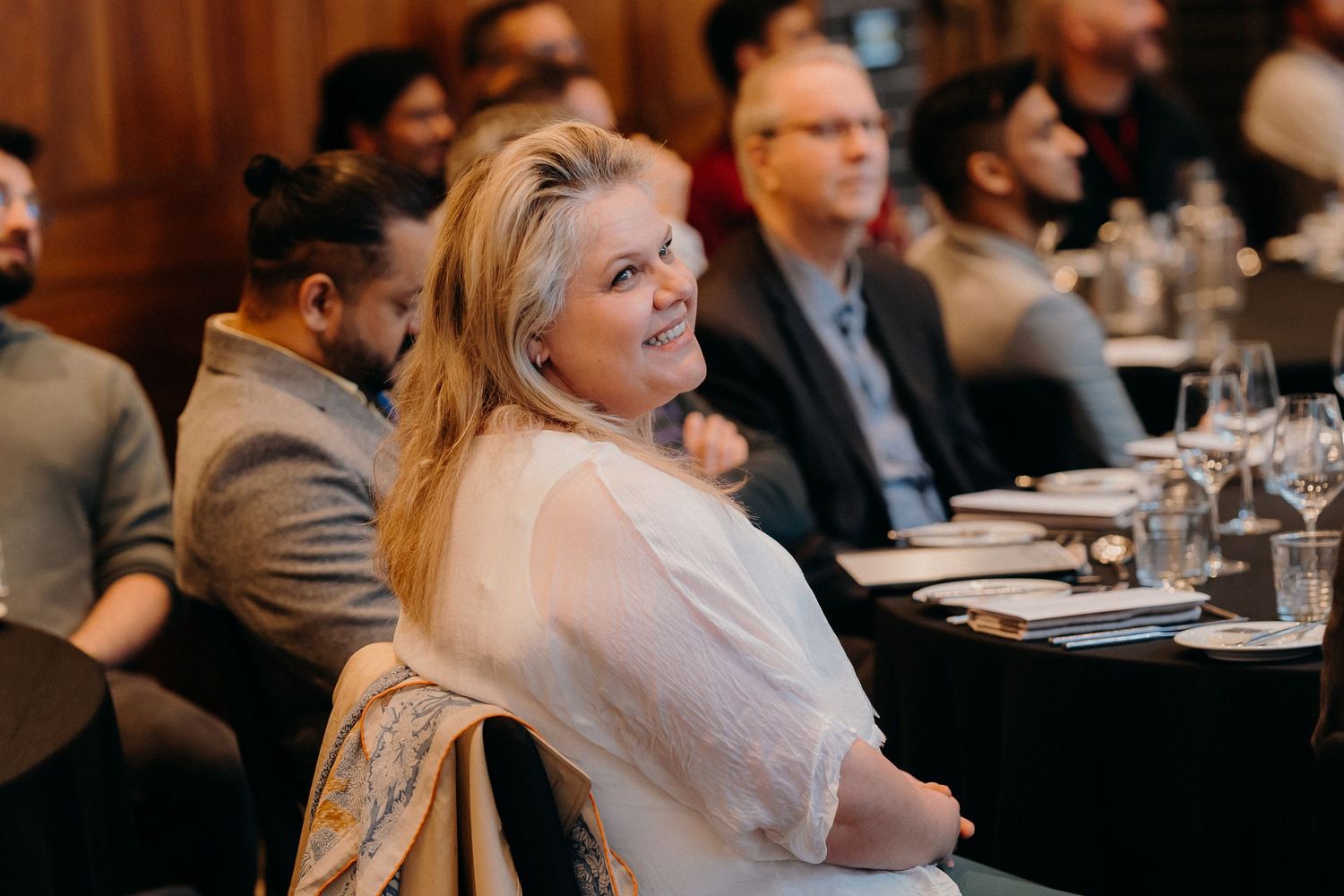 Business professionals seated at tables during a conference or seminar event in a dimly lit venue.