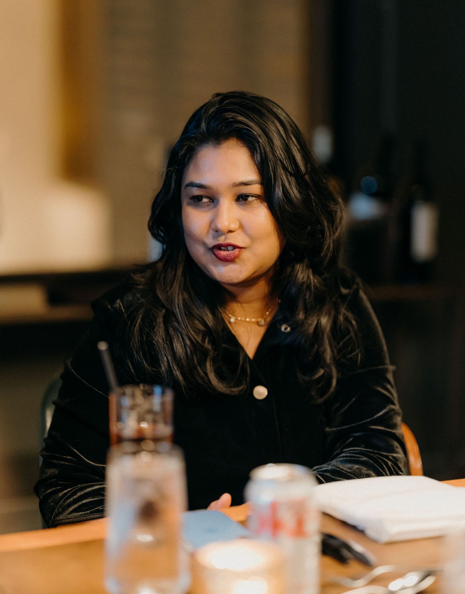 A person with dark hair sits at a table in a dimly lit restaurant setting.
