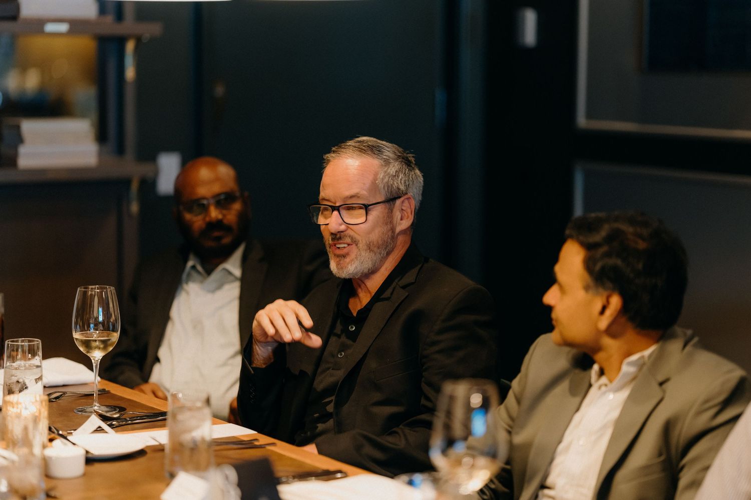 Business professionals engage in conversation during a dinner meeting at a dimly lit restaurant table.