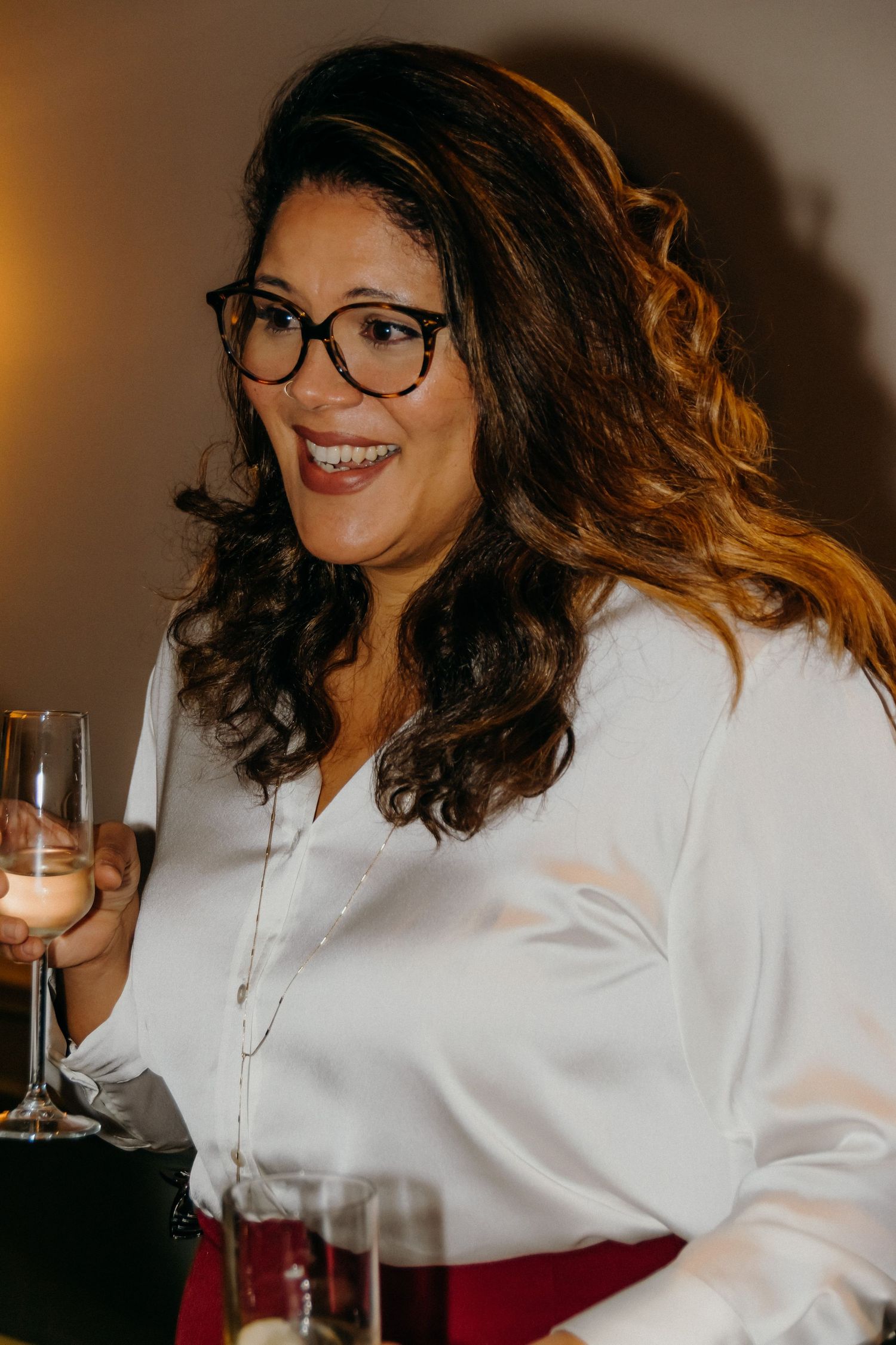Woman in white shirt and glasses smiling while holding wine glass in warm lighting.
