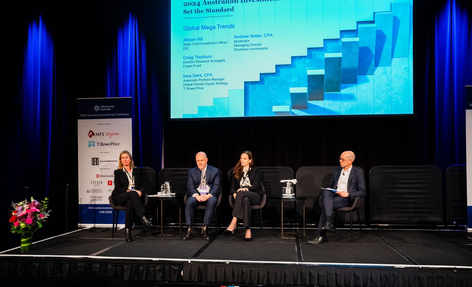 Business conference panel discussion with speakers seated on stage in front of large projection screens.