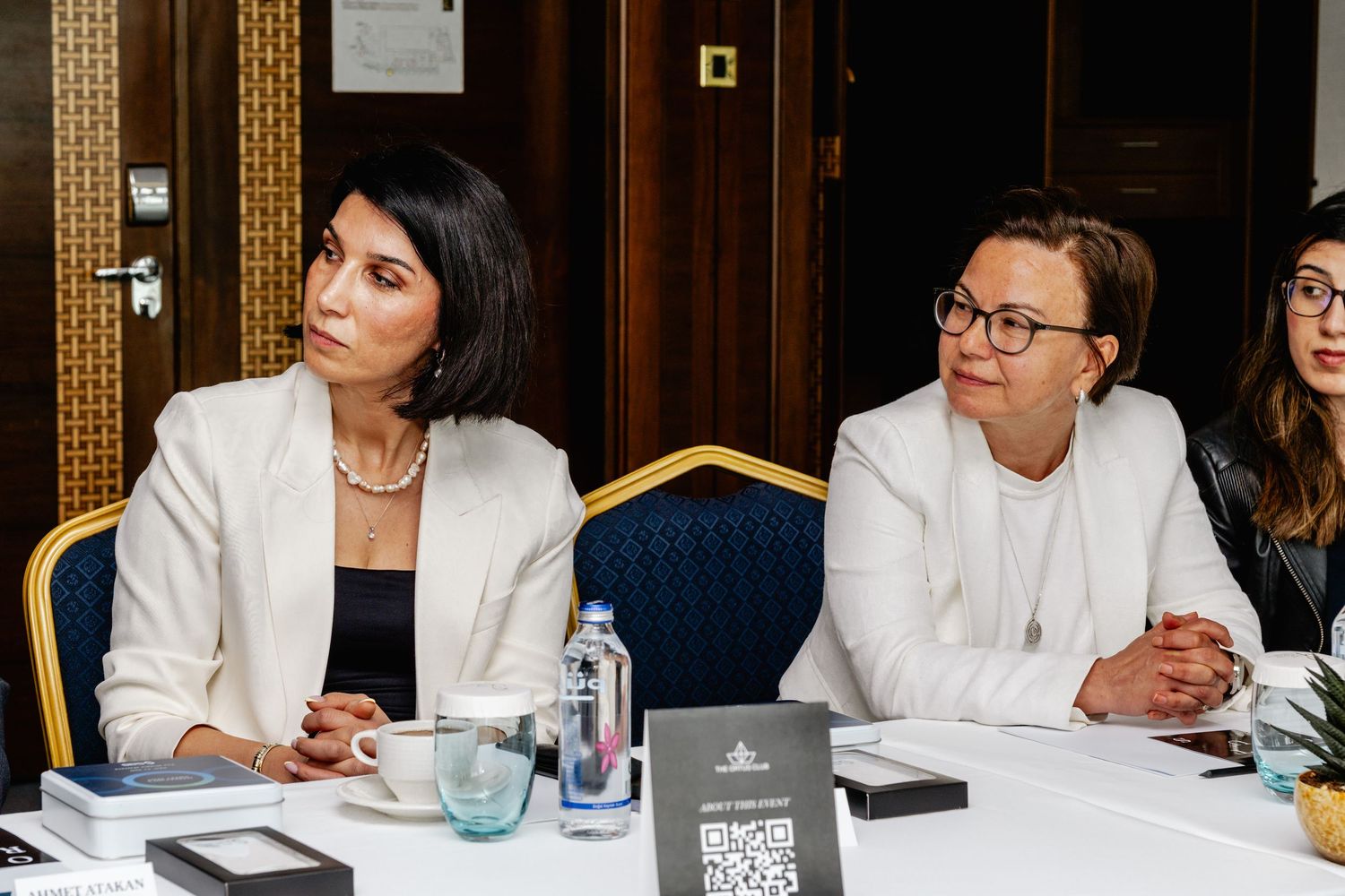 Two professional women in white blazers seated at a conference table during a formal business meeting.