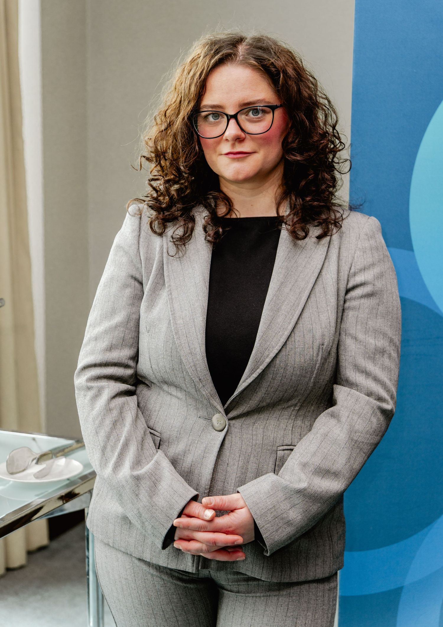 Professional woman in gray blazer and glasses with curly hair stands confidently in modern office setting.