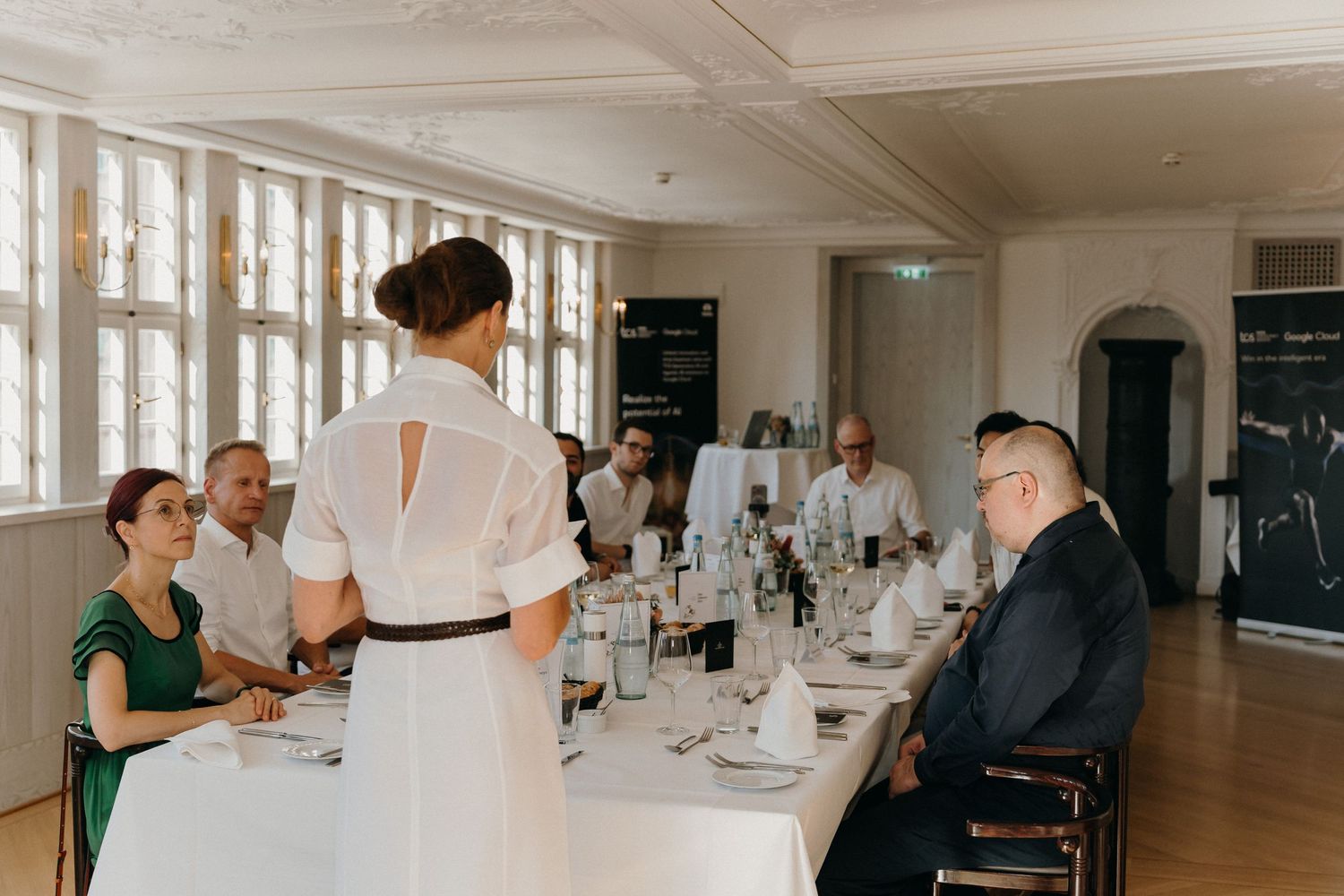 Elegant dinner event with guests seated at a long white-clothed table while a woman in white dress addresses the group.