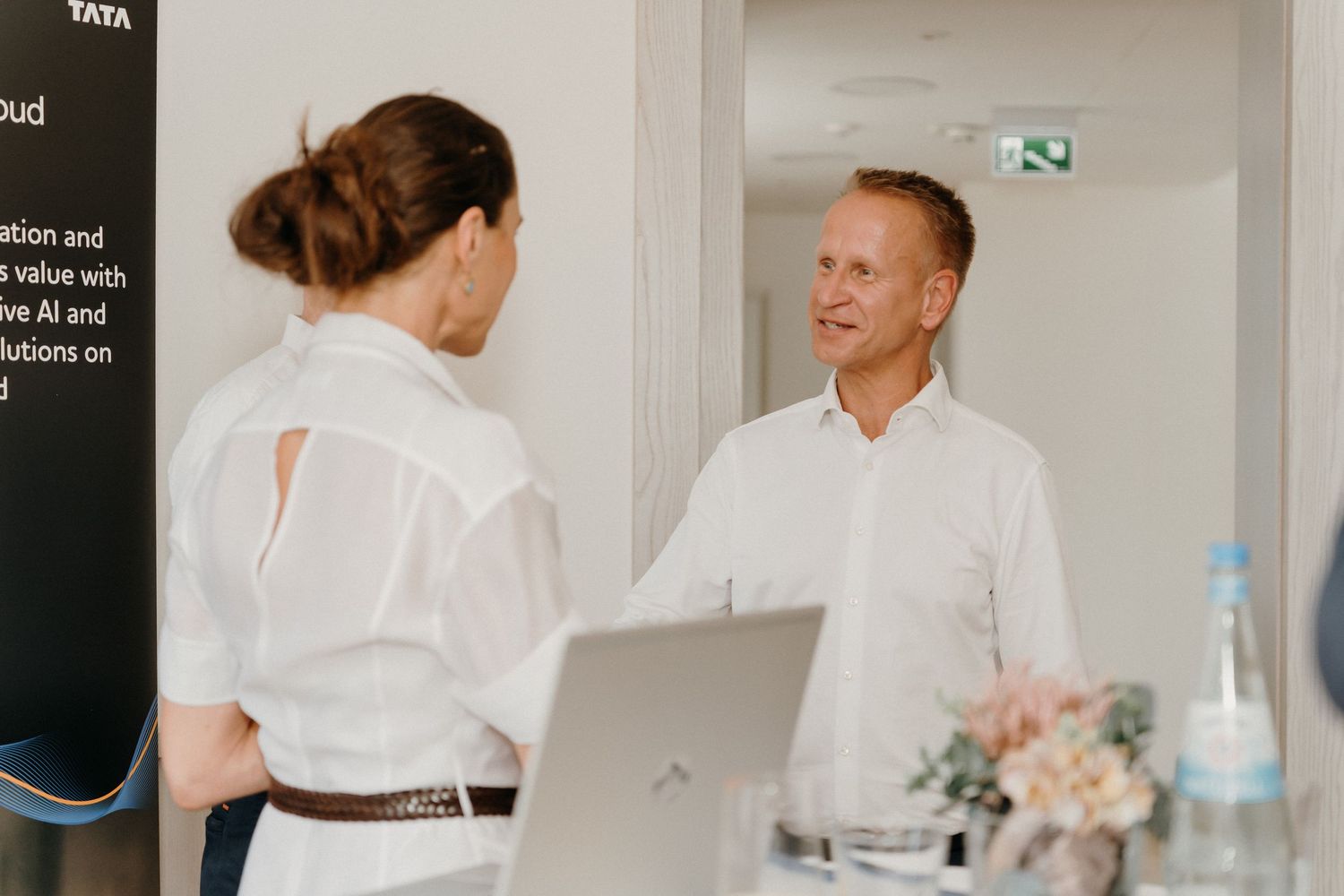 Two professionals in white shirts engaged in conversation at a Tata business event with AI branding visible.