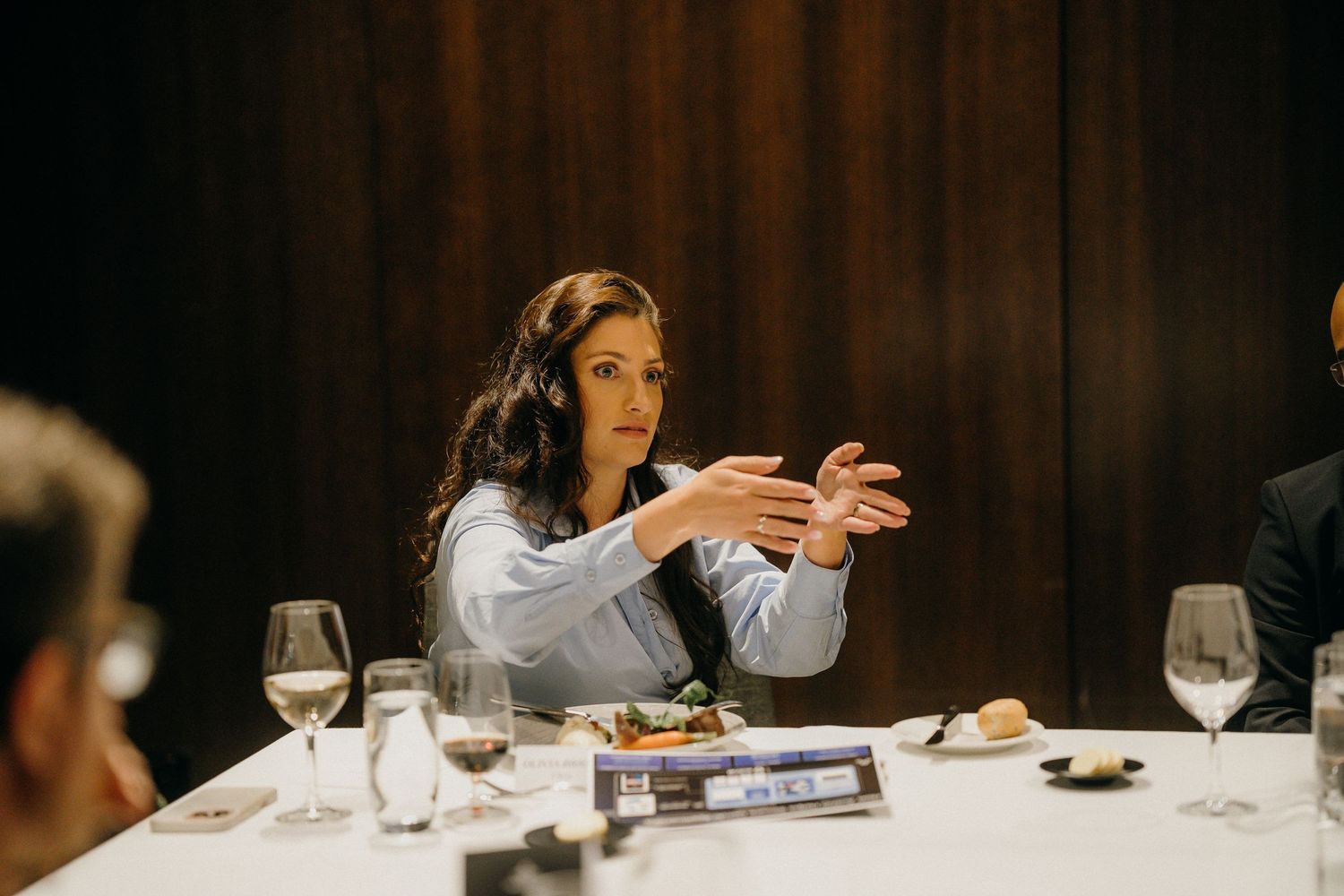 Woman gesturing expressively at an elegant dinner table with wine glasses and fine dining setting.