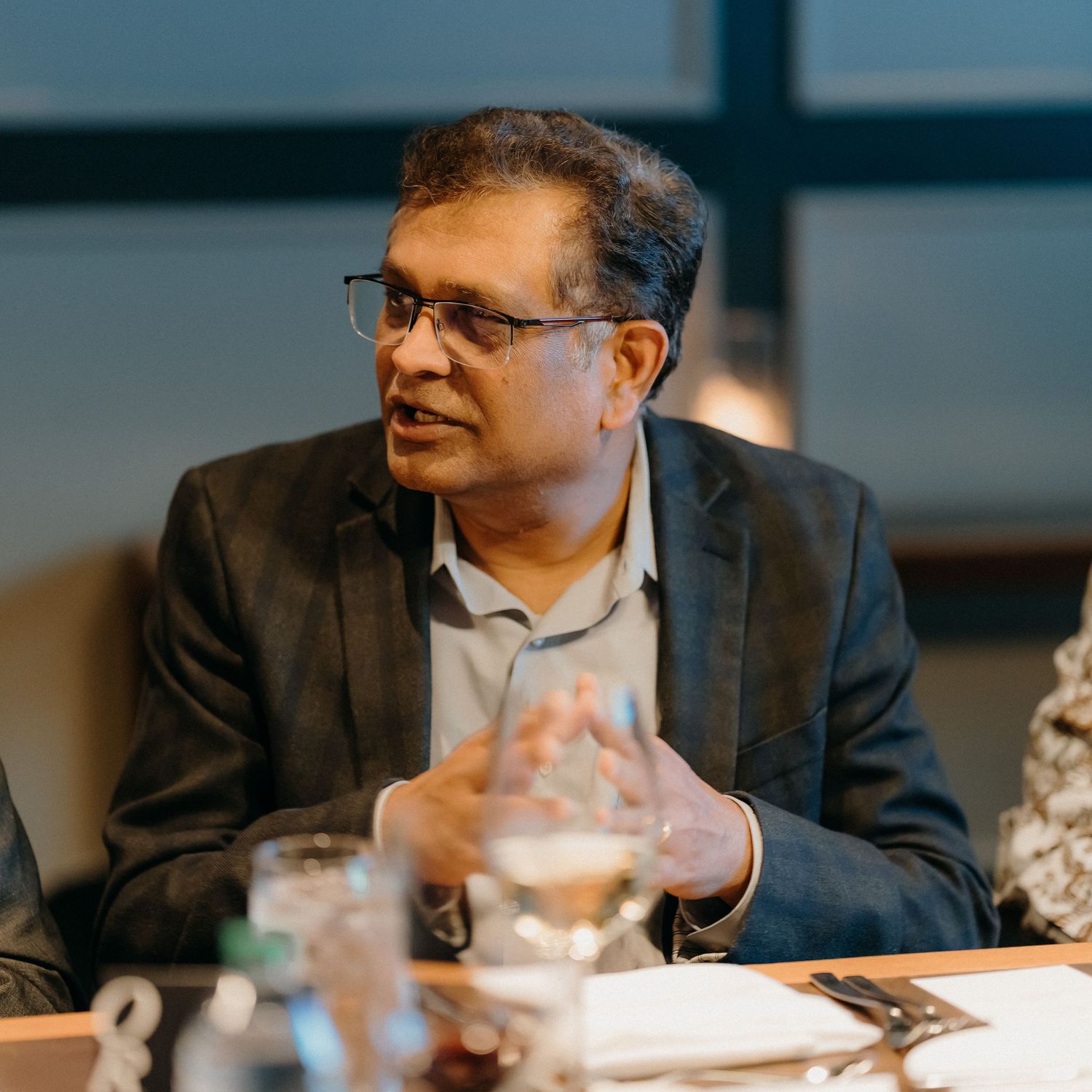 Professional in a dark suit jacket gestures while speaking during a business meeting at a conference table.