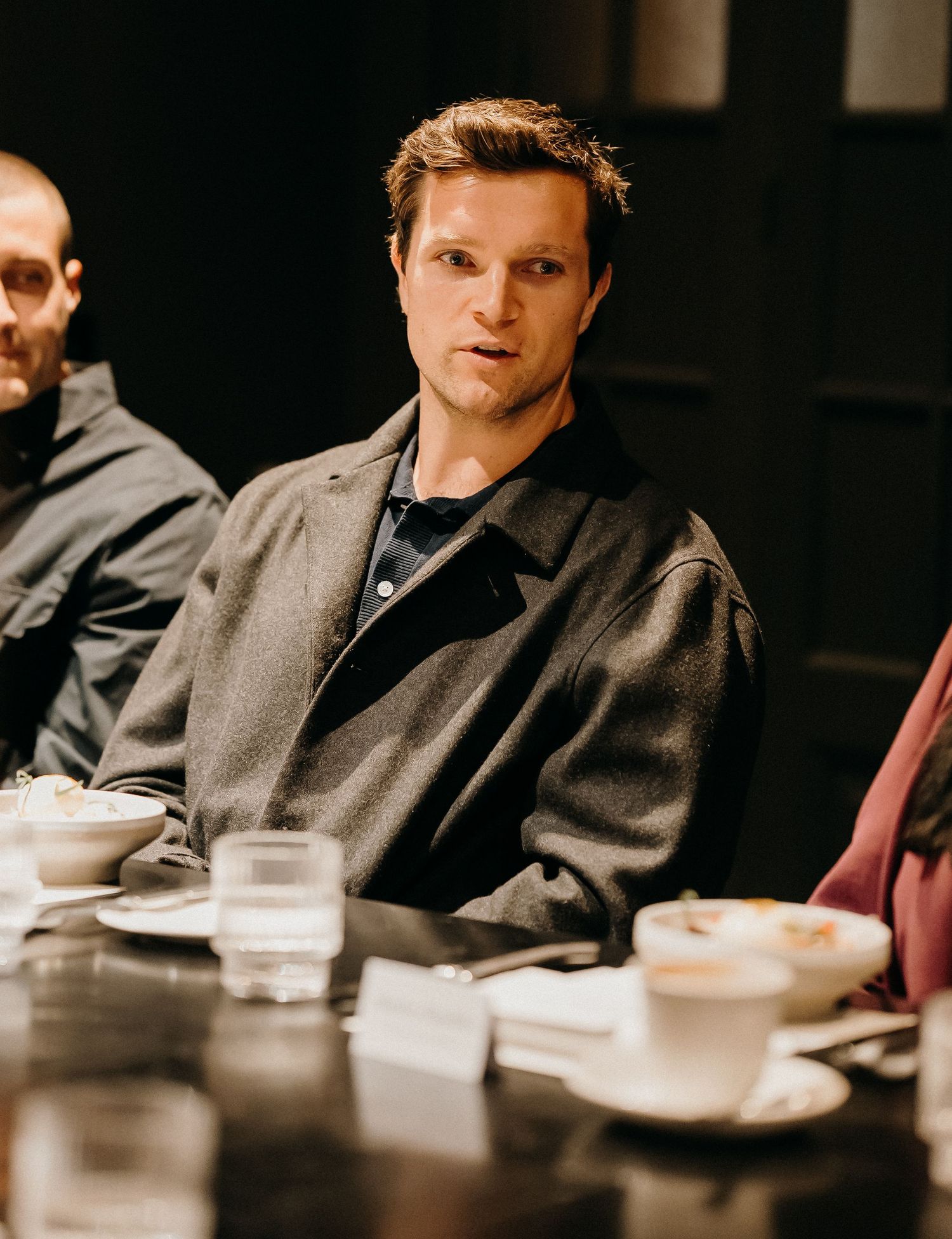A person in a dark suit sits at a dimly lit table with white dishes and cups in a restaurant setting.