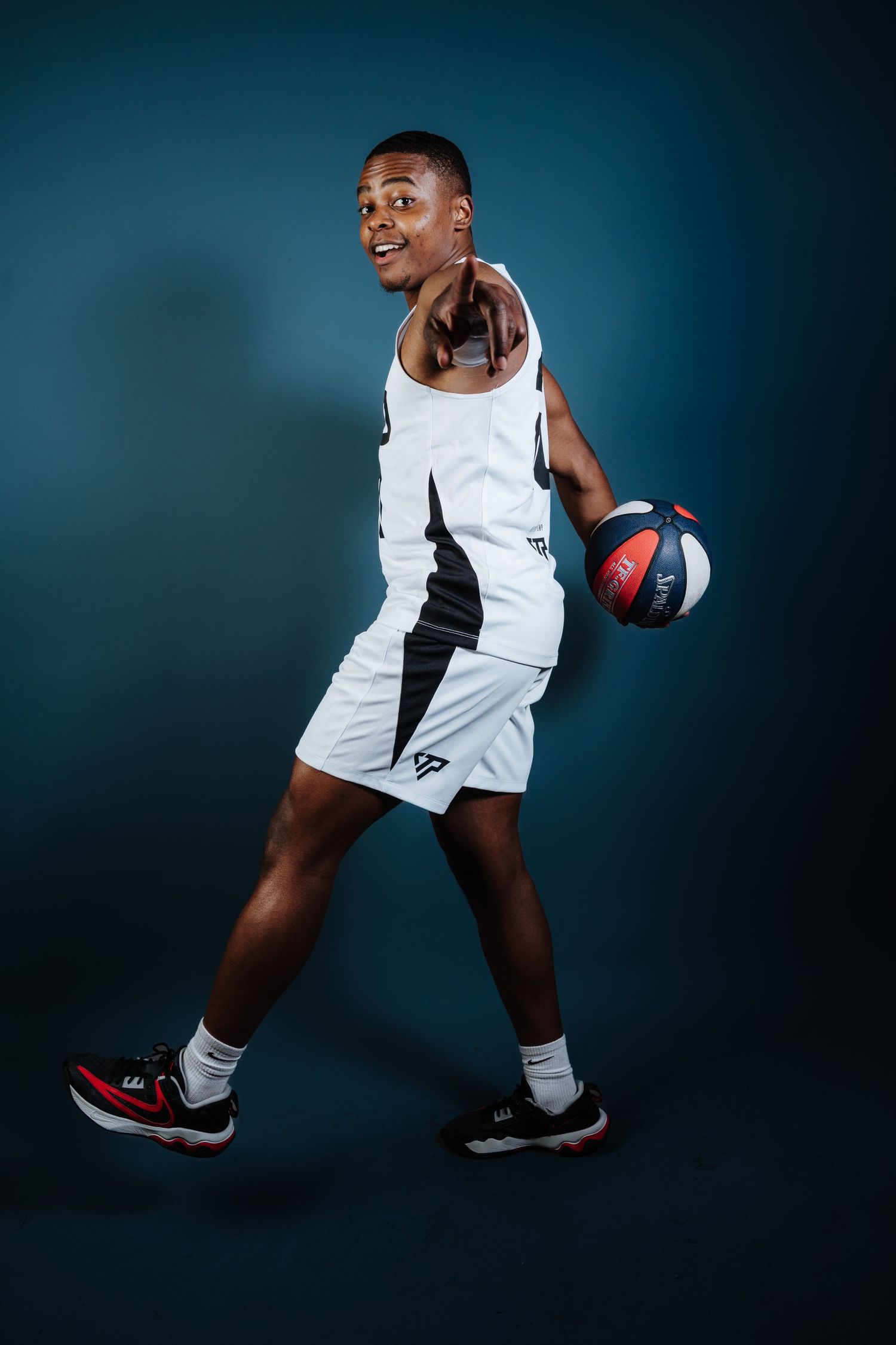 Basketball player in white uniform poses with ball against dark blue background during studio photoshoot.
