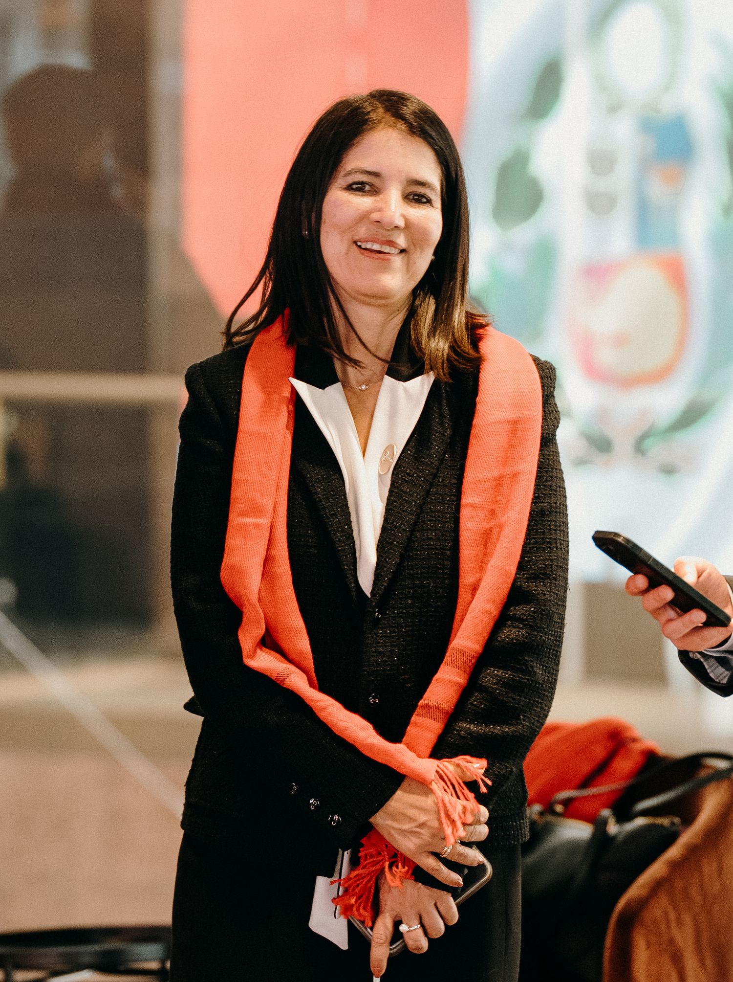 A person in a black blazer and orange scarf smiles while seated during what appears to be a professional event.