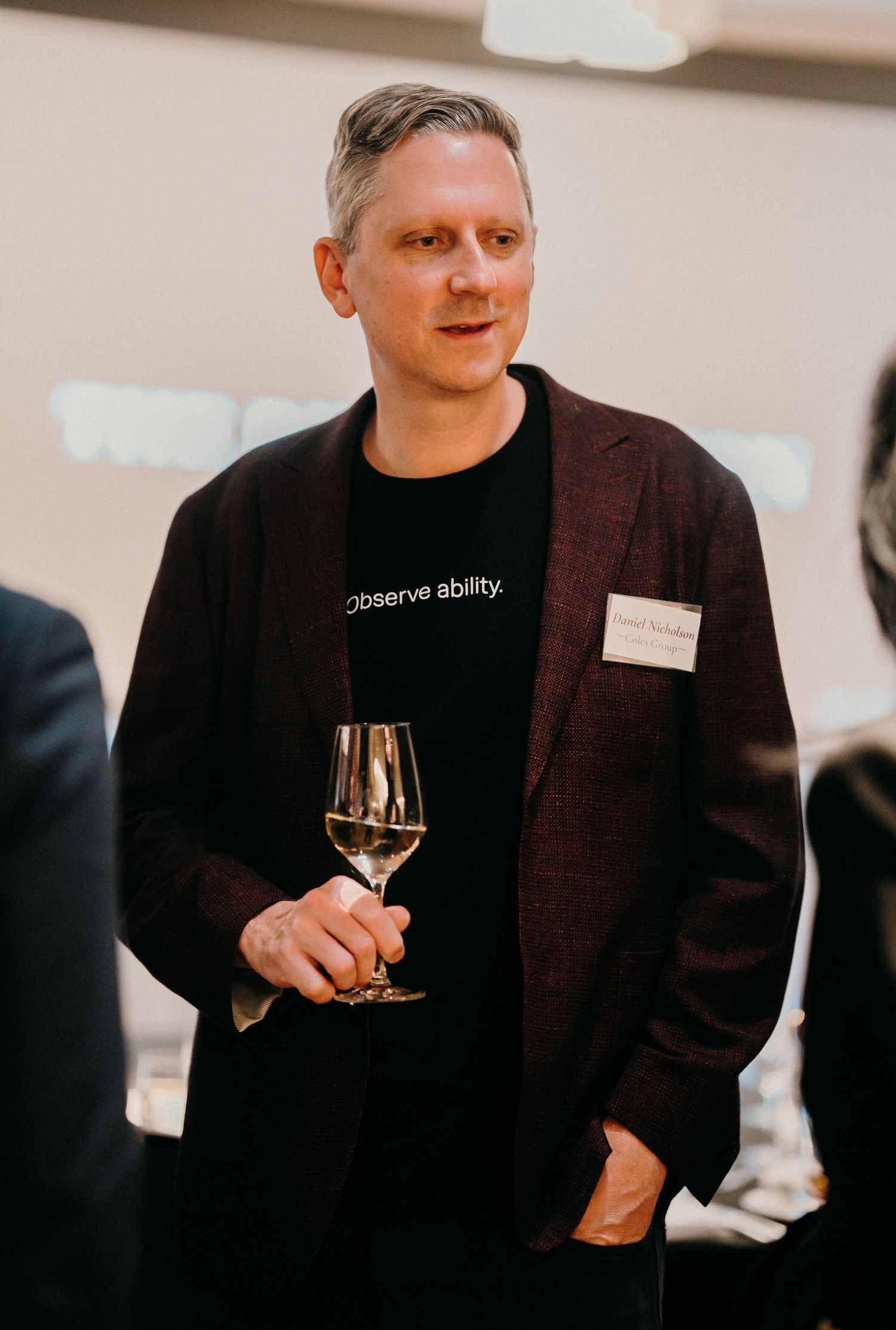 Person in a black jacket and shirt holding a glass of white wine at an event.