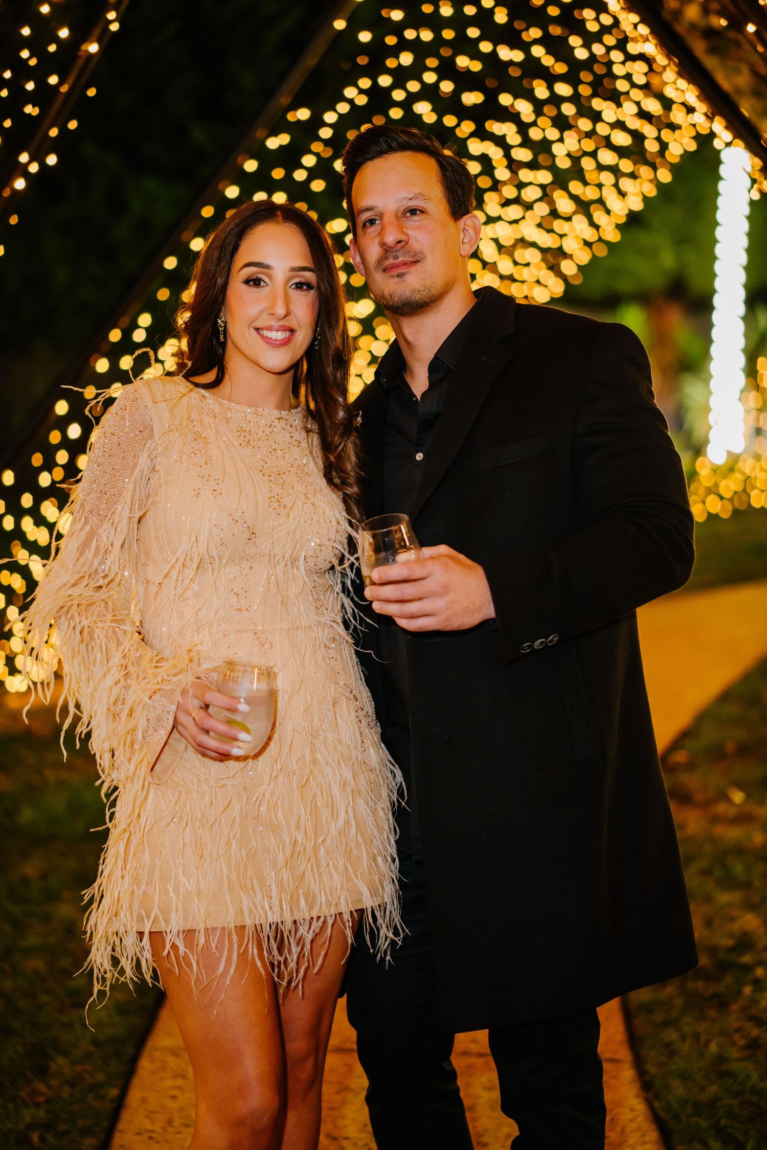 A couple poses together at night in front of glowing string lights, with one wearing a beige feathered dress and one in black.