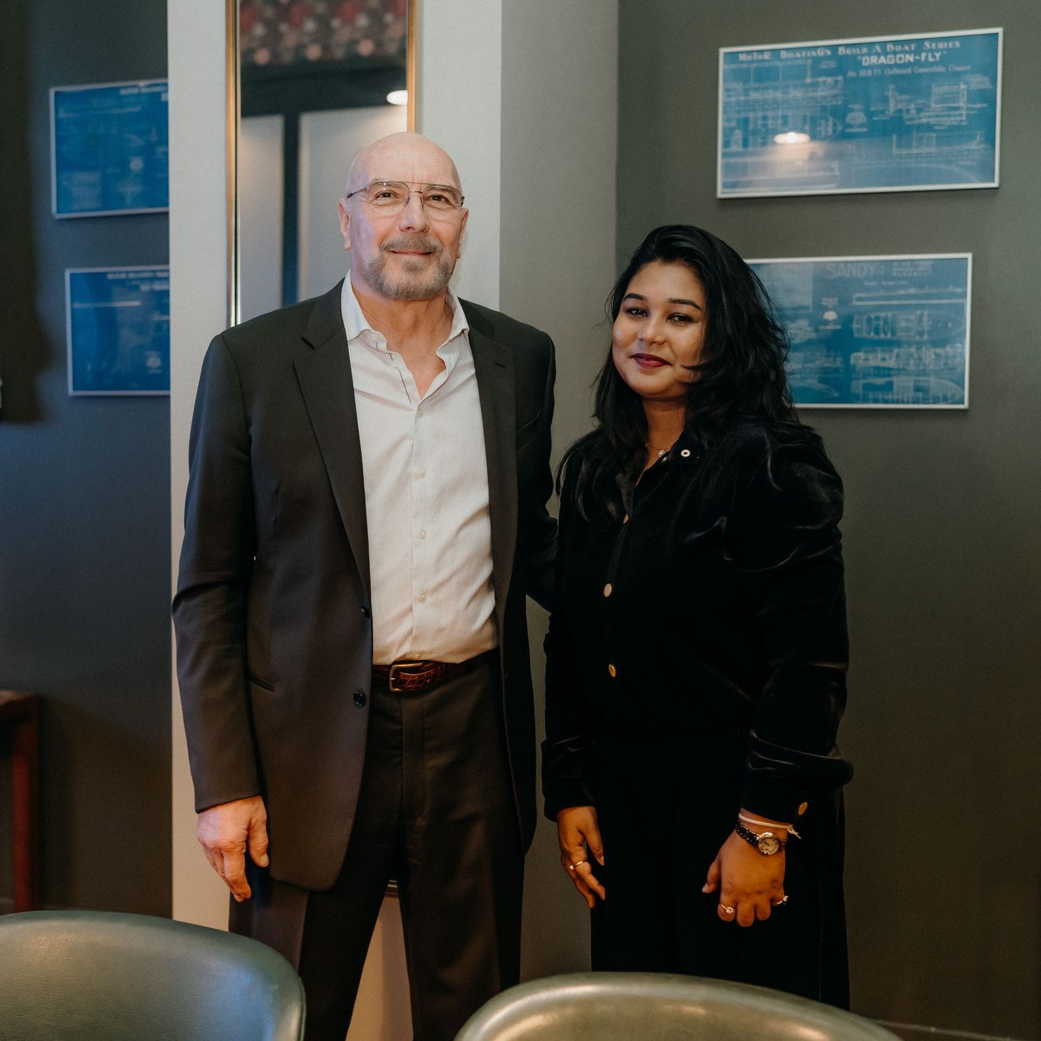 Two people in business attire standing and posing together in an office hallway with blueprints visible on the wall.