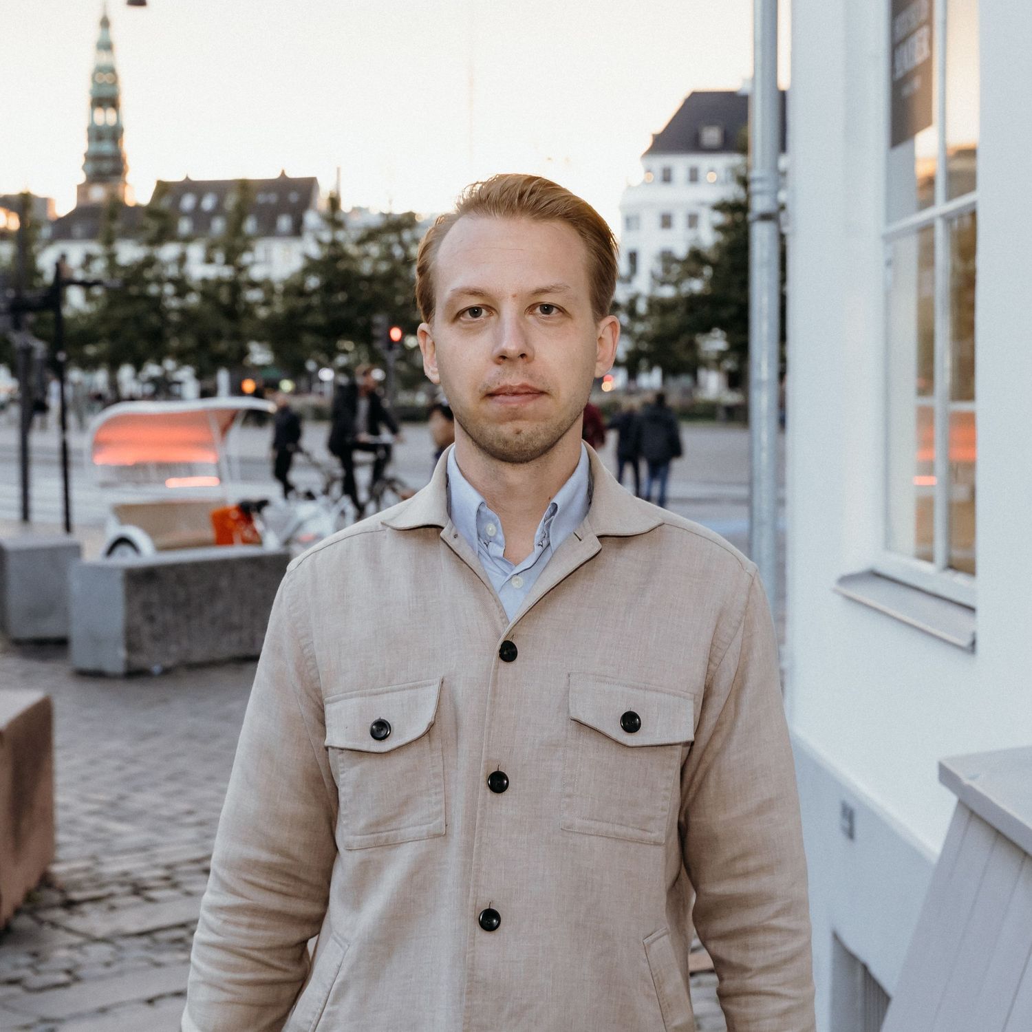 Professional portrait of someone wearing a beige coat and light shirt standing against a white building exterior.