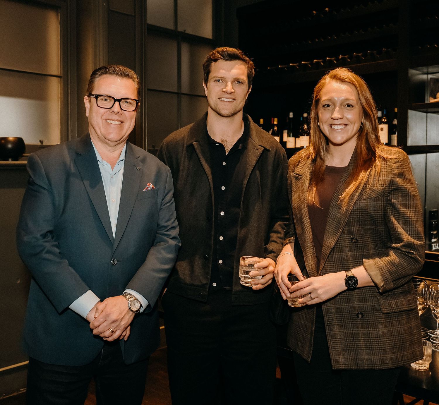 Three people pose together at an event, with one wearing a navy suit and glasses, and another in a plaid blazer.