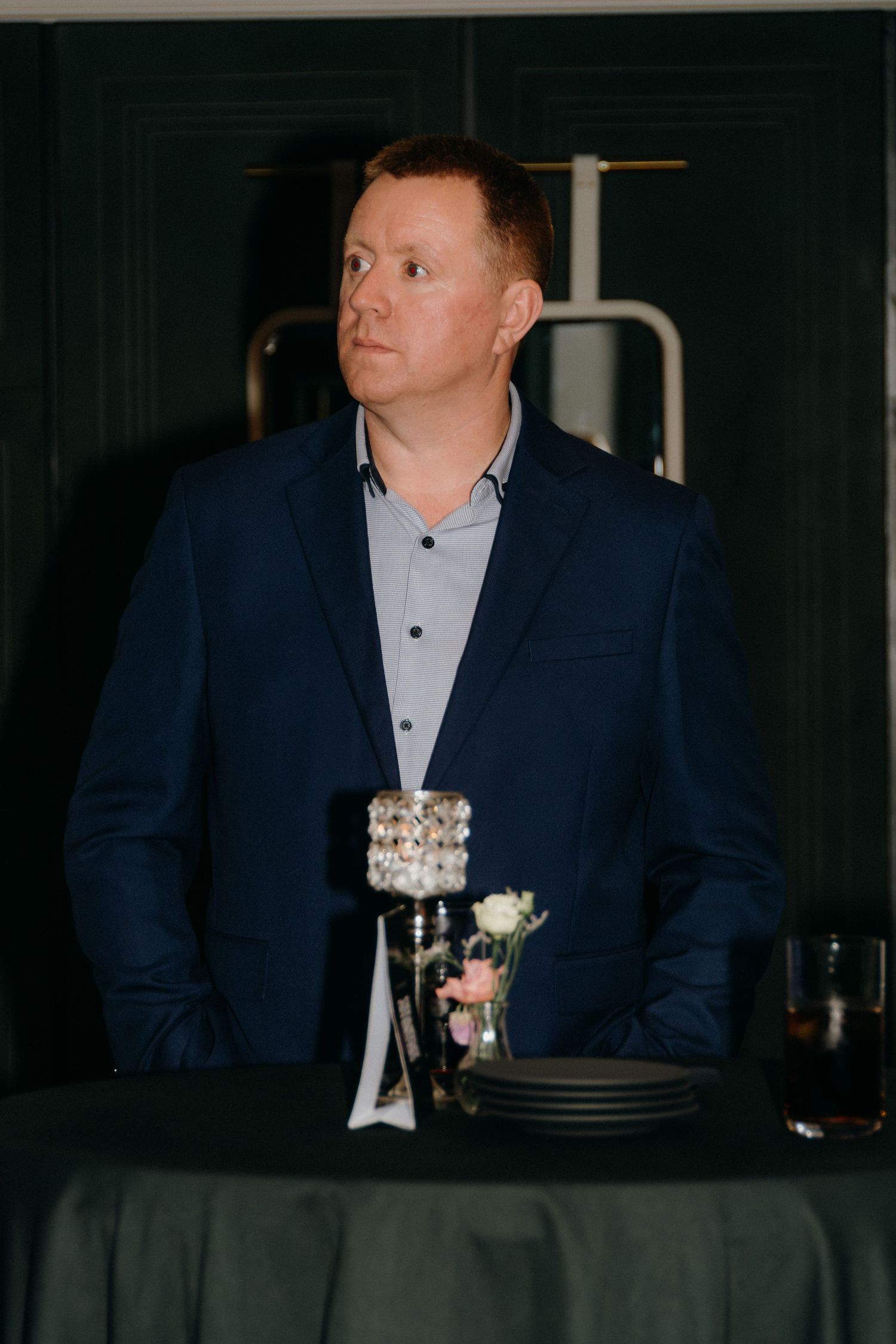 A person in a navy blue suit sits at a table with a glass of wine in a dimly lit setting.
