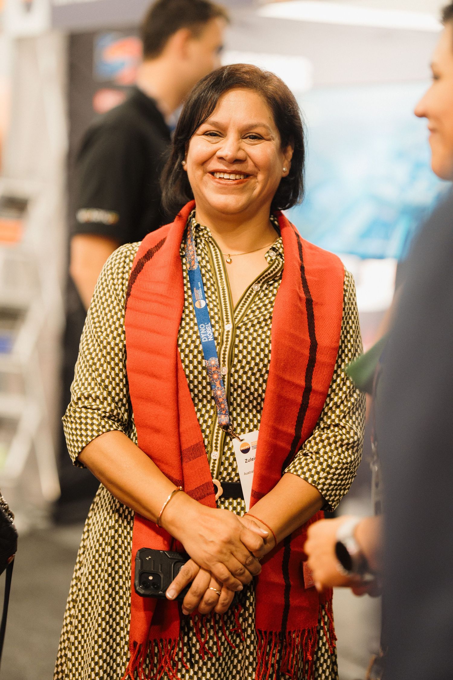 A person wearing a red vest over a patterned shirt smiles warmly at a professional event.