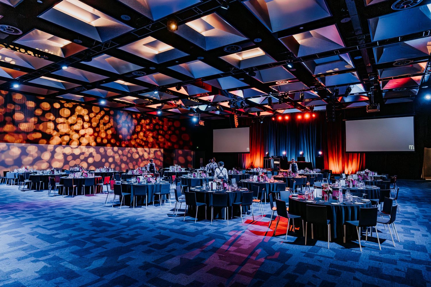 Modern event venue with geometric ceiling panels, illuminated in blue and red lighting over round dining tables.