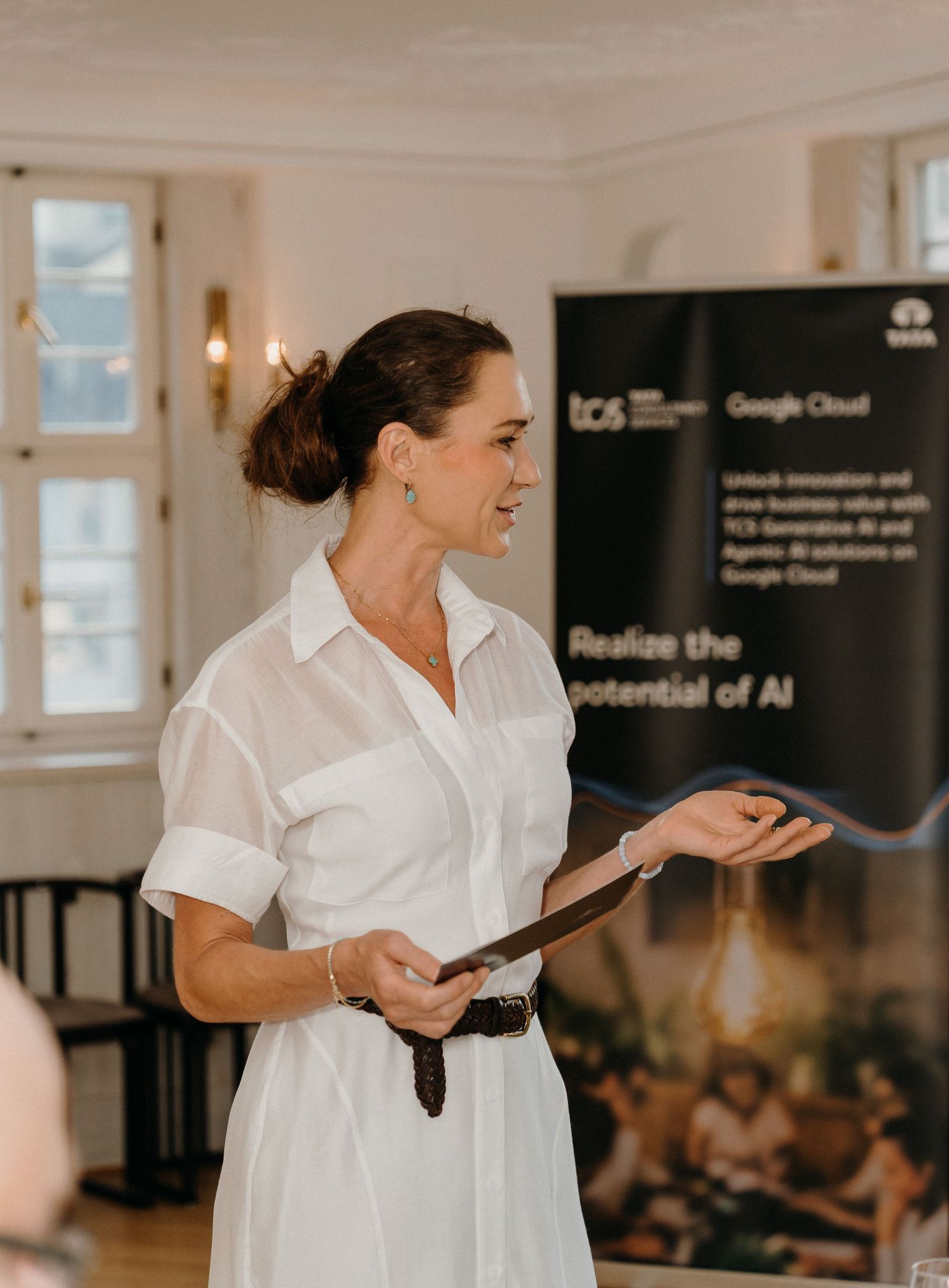 Woman in white dress presenting at TCS and Google Cloud AI event, holding tablet while speaking to audience.