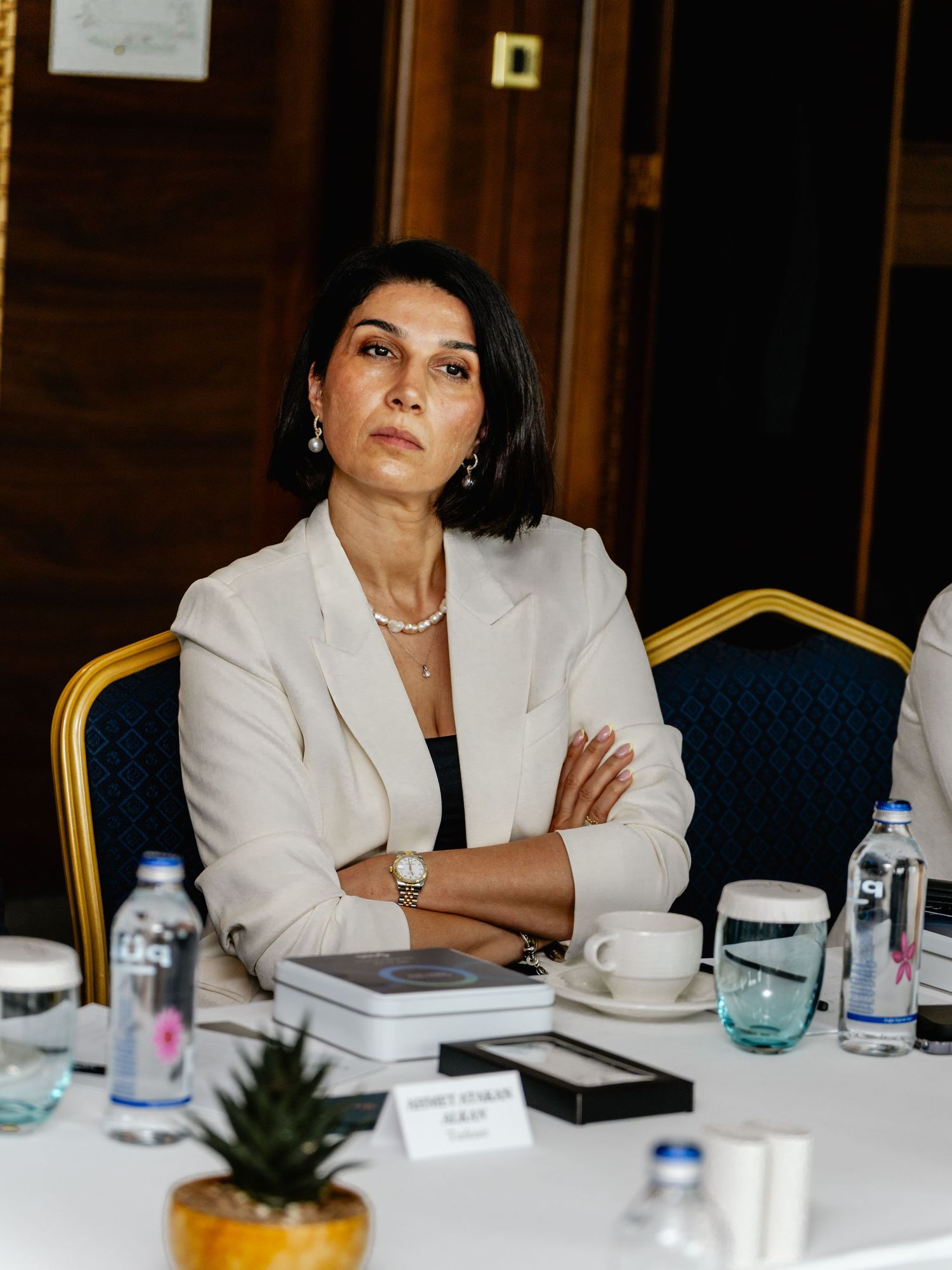Professional woman in white blazer sitting at conference table with arms crossed, looking attentive.