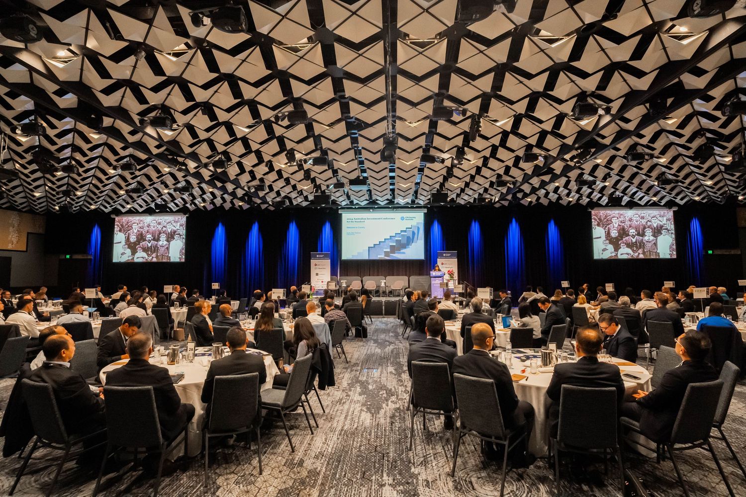 Large conference hall with geometric ceiling pattern and rows of tables set for a formal event.