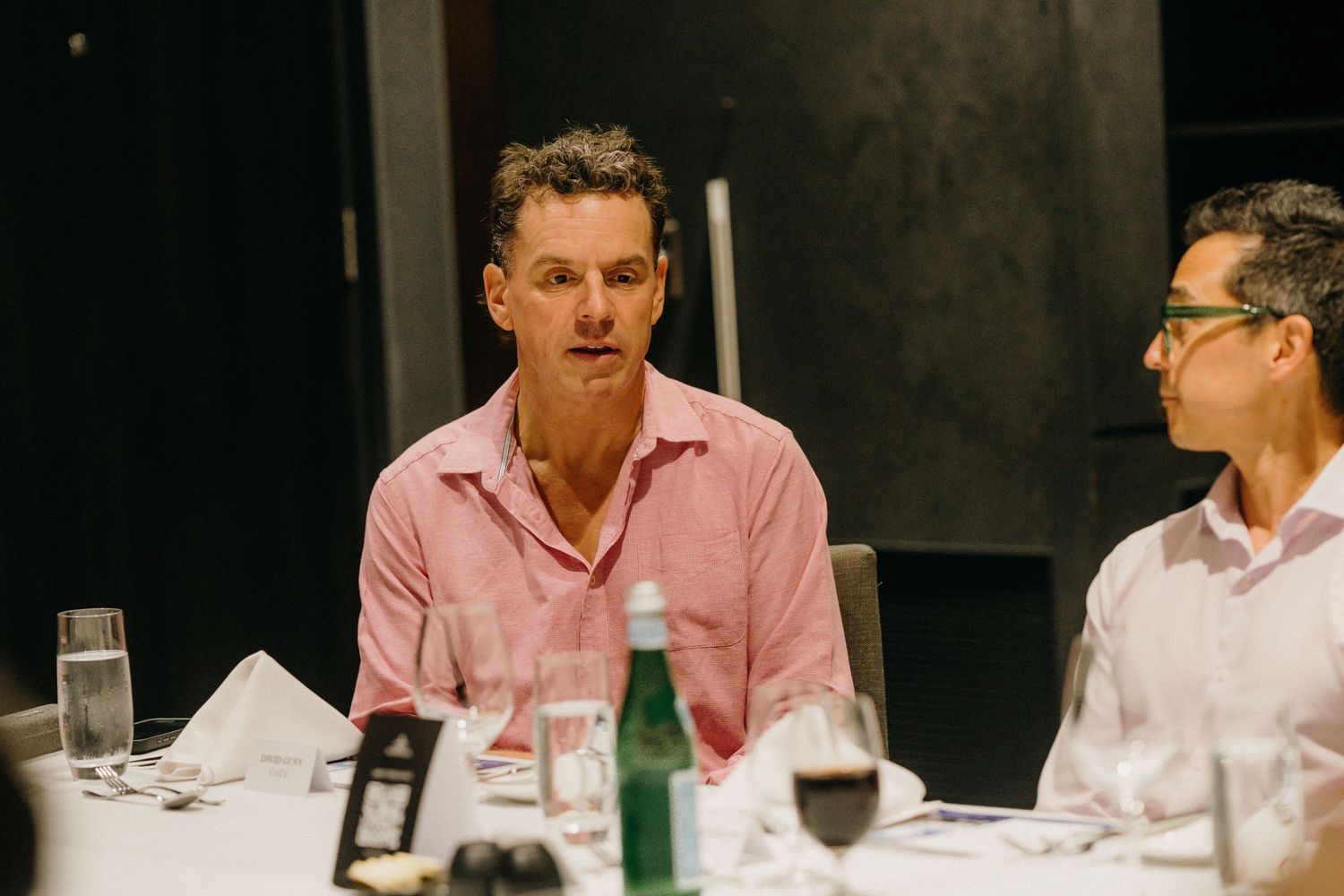 Two men in conversation at a formal dinner table with wine glasses and water bottles.