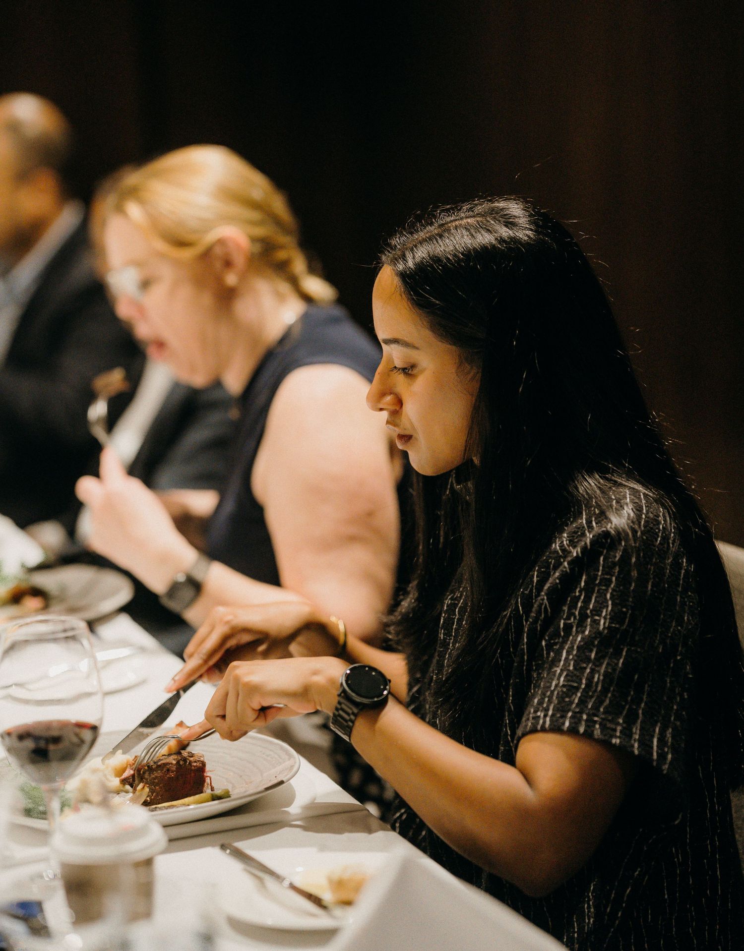 Guests dining at a formal event, with a woman in black cutting food at an elegantly set table.