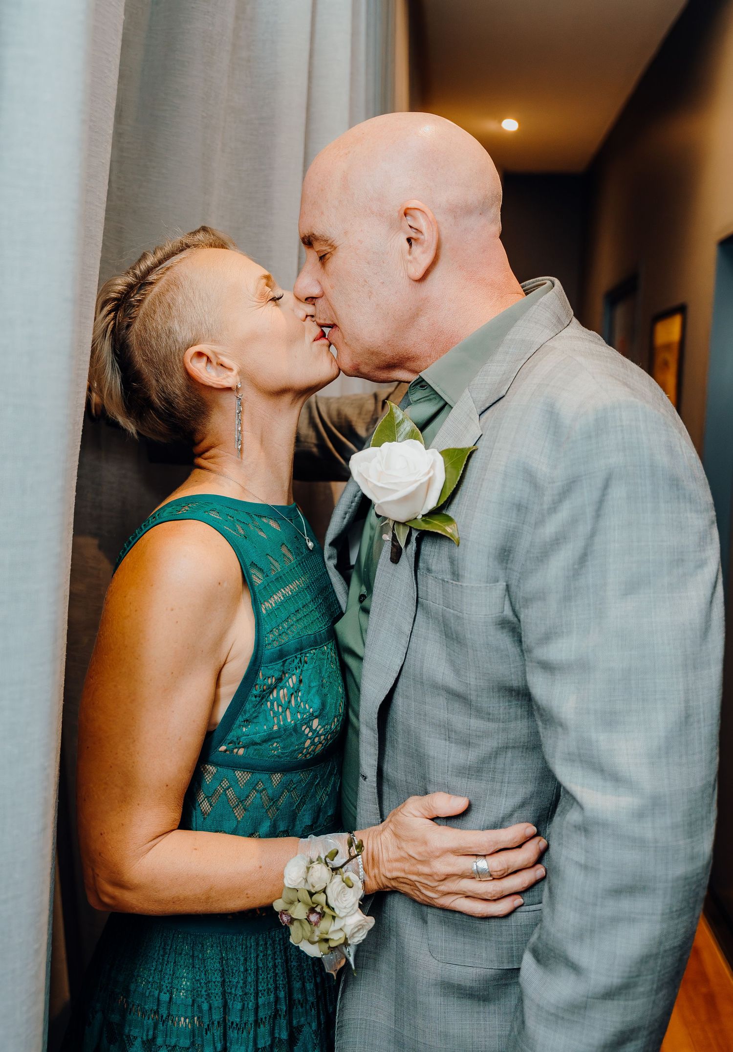 A couple in a gray suit and green dress share an intimate kiss against a backdrop of white columns.
