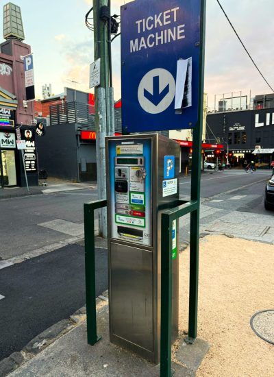Multiple parking meter ticket machines installed along an urban sidewalk with blue signage visible in the background.