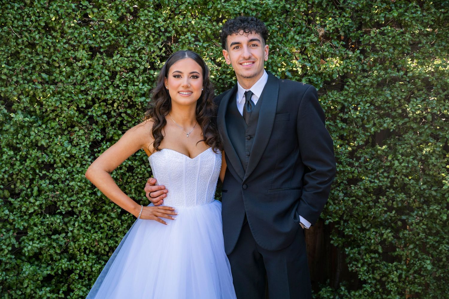 A happy couple poses for wedding photos outdoors with a lush green hedge backdrop.