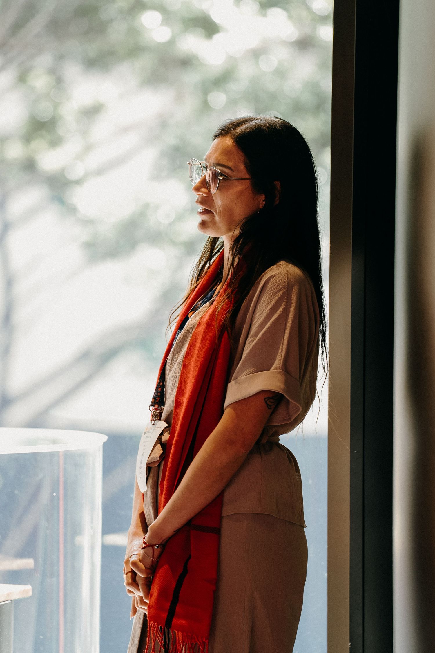 A person wearing glasses and a red scarf stands by a window, looking up with a contemplative expression.