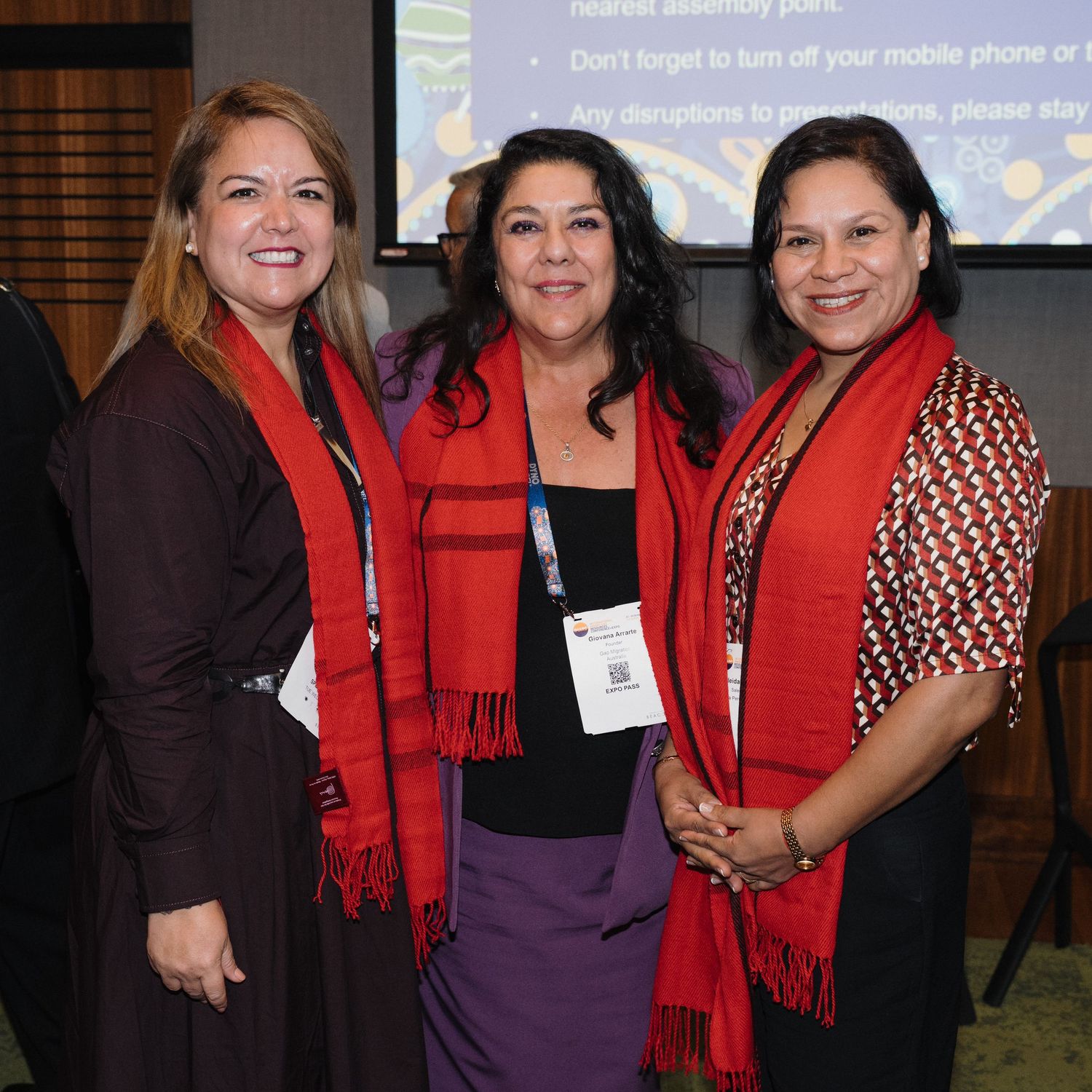 Three women wearing red scarves and professional attire pose together at a business event.