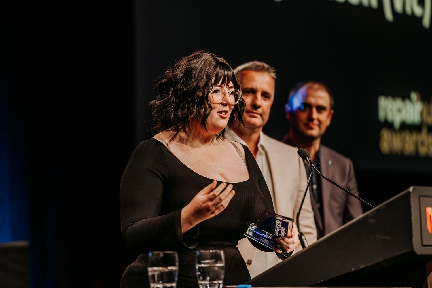 Speakers at a professional conference engage in discussion while standing at podiums on a dark stage.