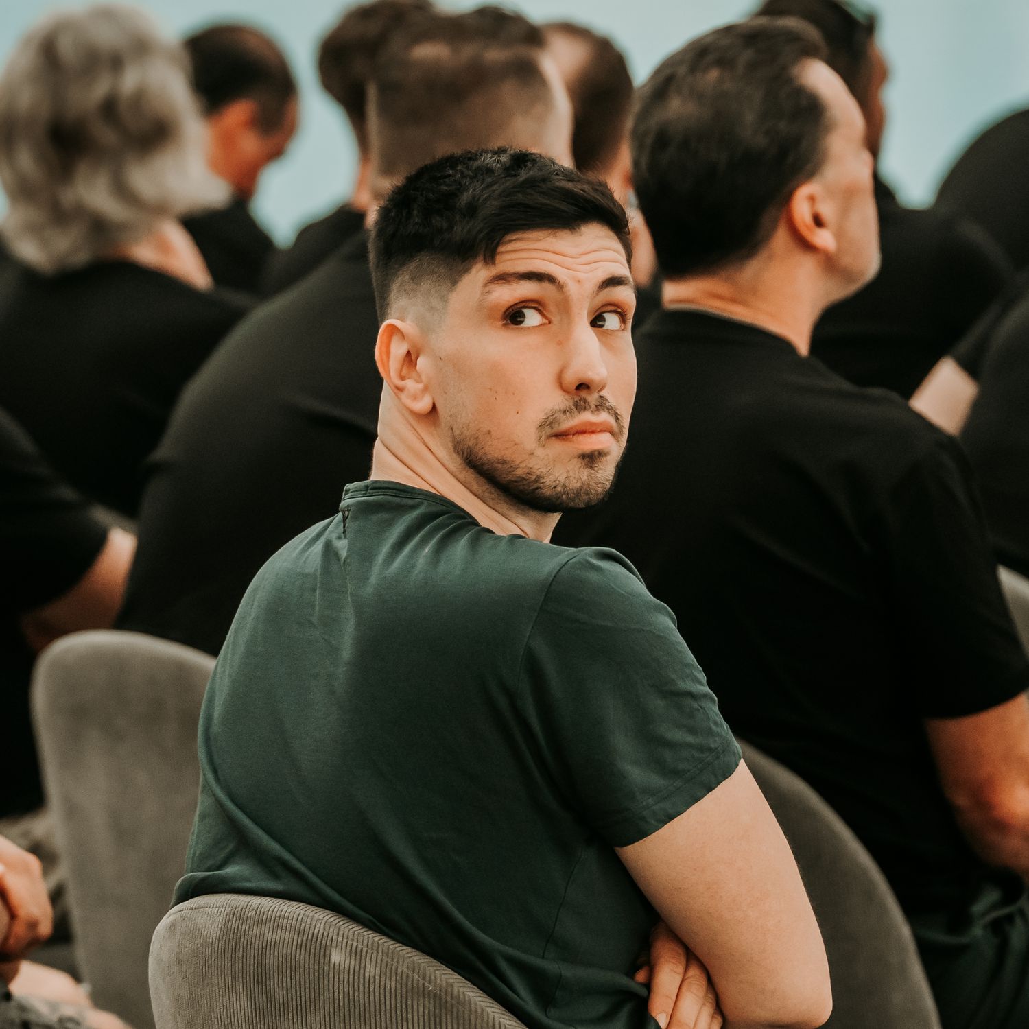 A person in a black t-shirt sits in a dimly lit room among a crowd looking upward thoughtfully.