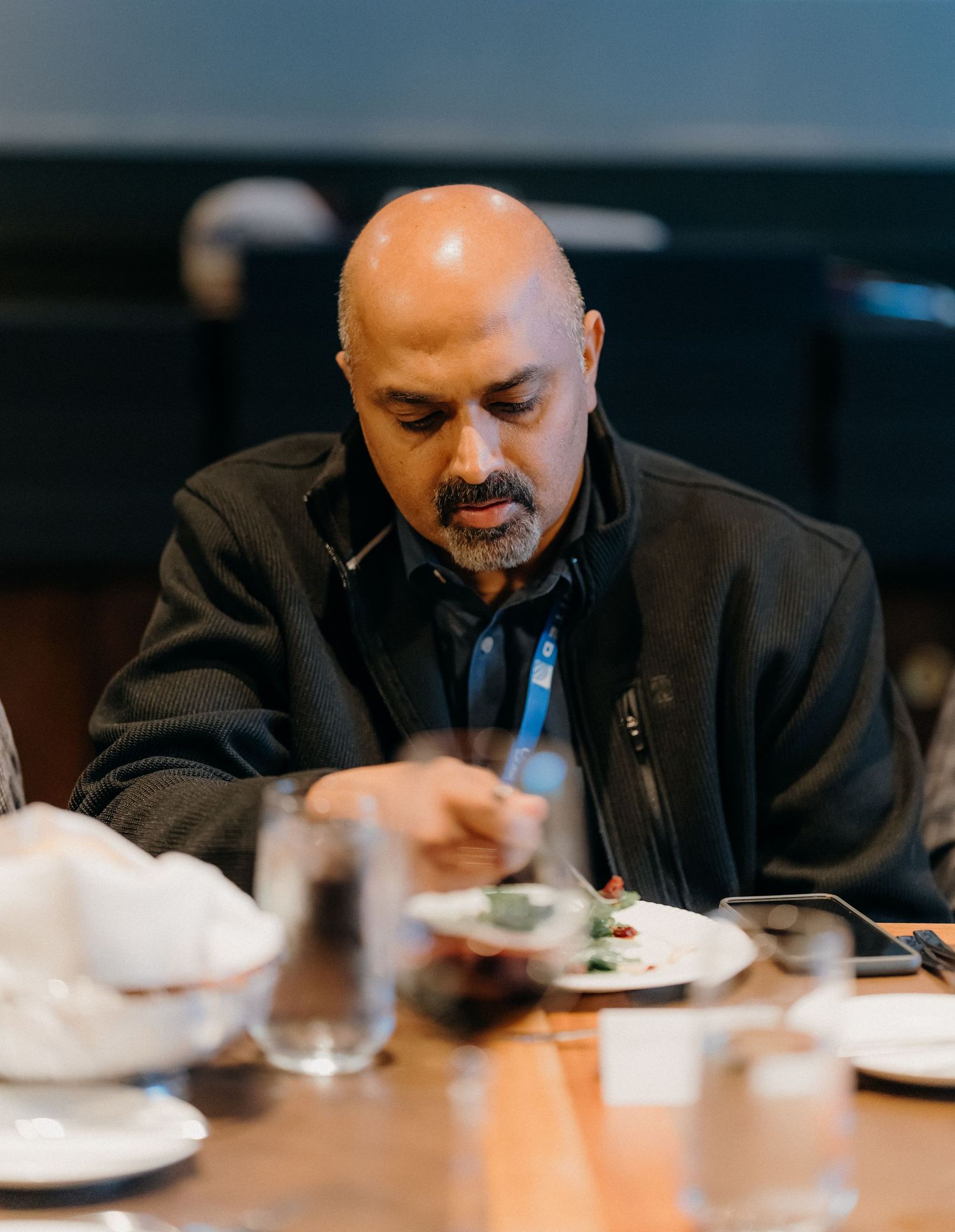 A person in a black jacket sits at a dining table looking down at their meal.