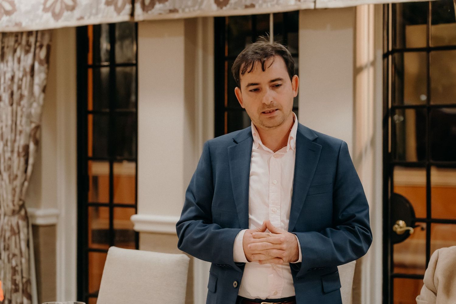 A person in a navy suit stands in front of a white wall with windows, creating a professional business portrait.