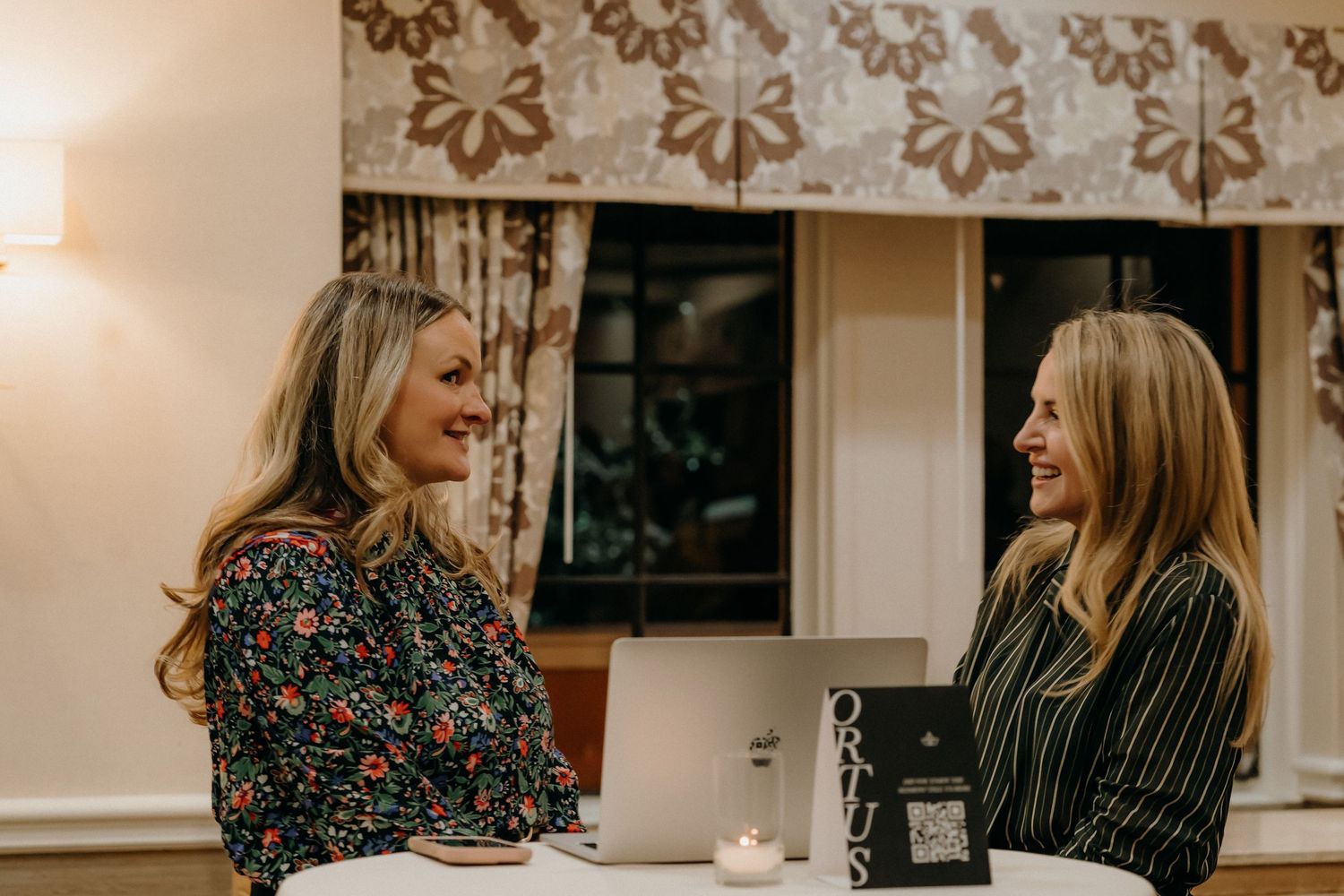 Two people sit at a table having an animated conversation in a room with floral wallpaper and warm lighting.