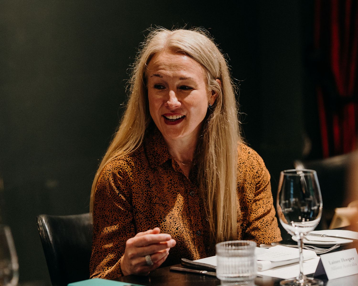 Woman in brown sweater laughs while dining at restaurant table with wine glass and place settings.
