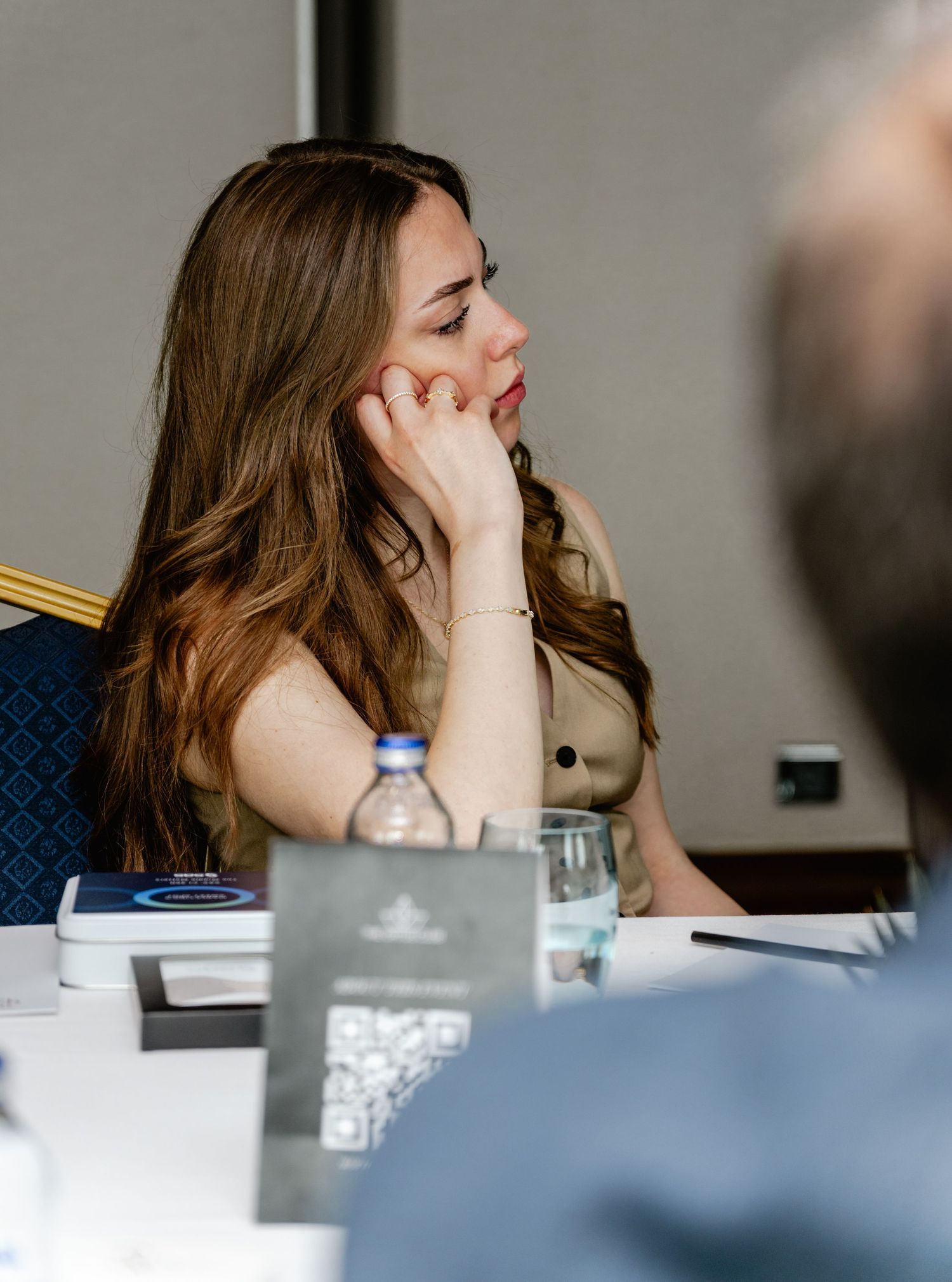 Woman with long brown hair sitting at a conference table, resting her chin on her hand in a thoughtful pose.