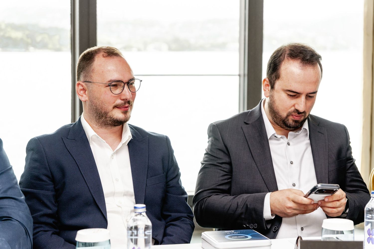 Two businessmen in suits sitting at a conference table, one checking his phone during a corporate meeting.