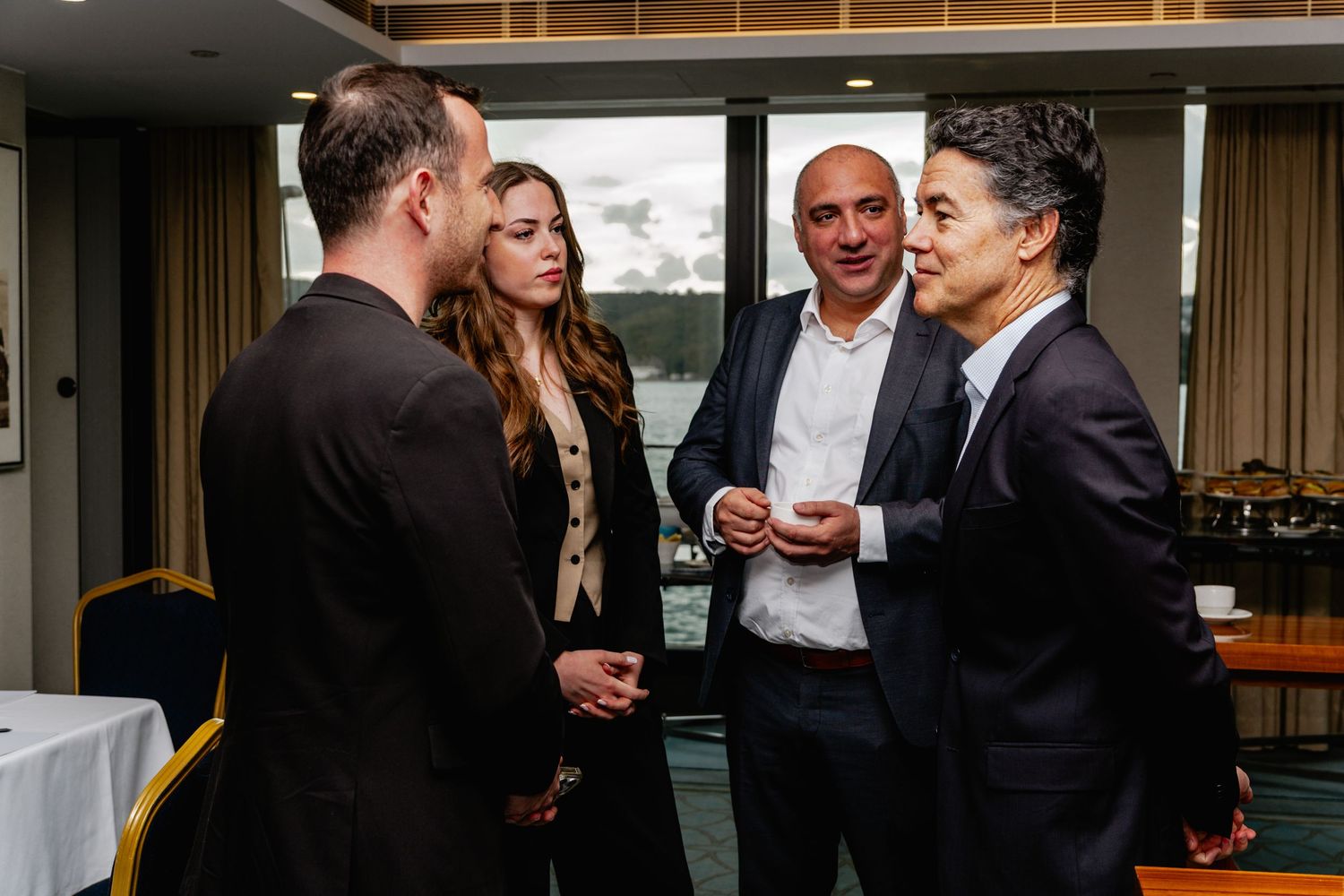 Four professionals in formal attire engaged in conversation at a corporate networking event with harbor views.