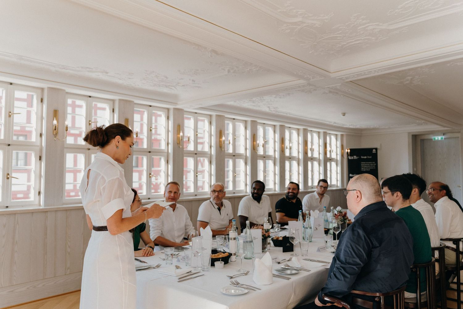 Woman in white presenting to guests at an elegant dining table in a bright, upscale restaurant setting.