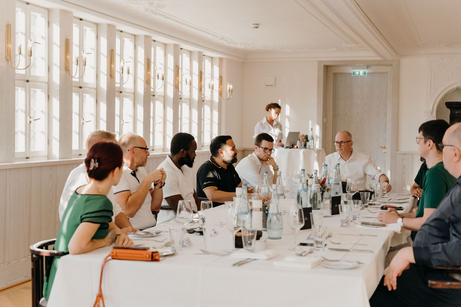 Elegant dinner party gathering around a long white-clothed table in a bright dining room with guests engaged in conversation.