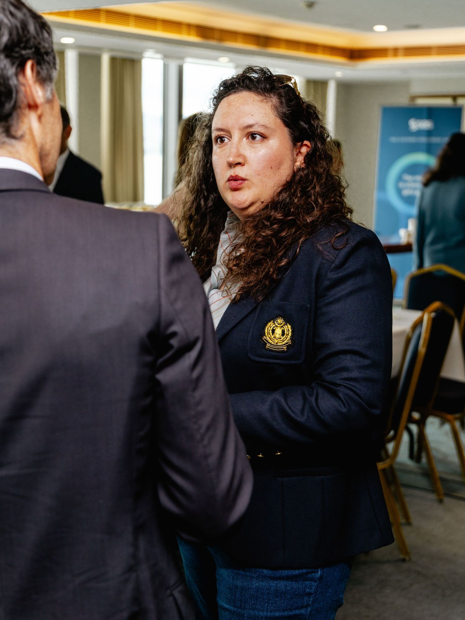 Woman in navy blazer with gold emblem engages in conversation at a professional networking event.