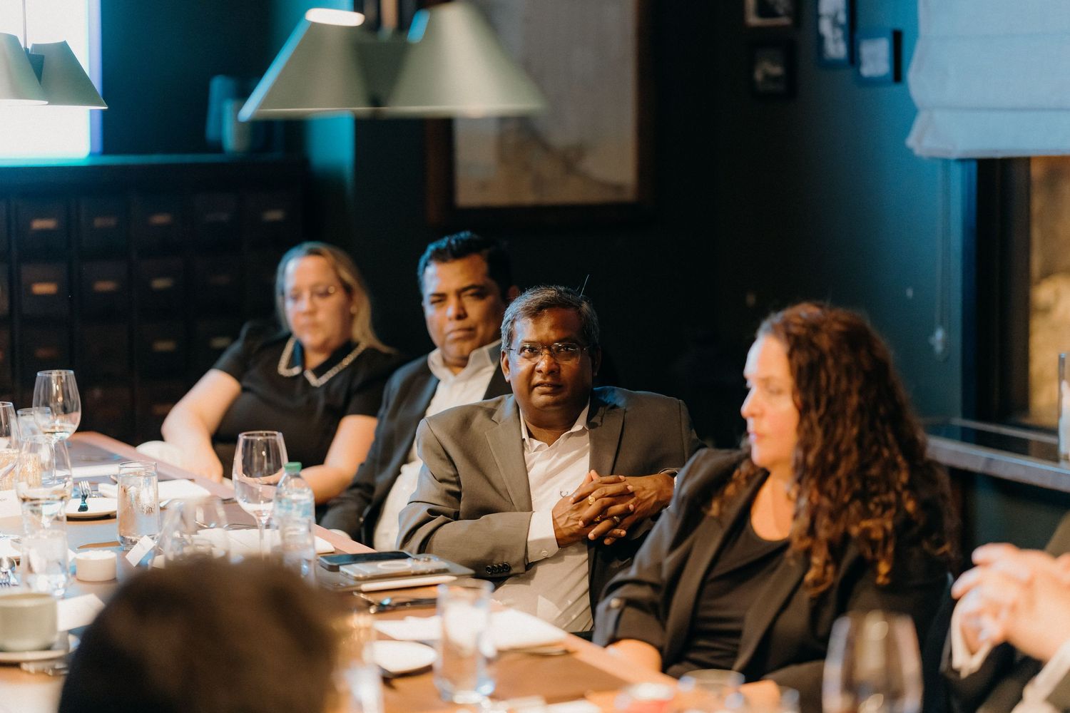 A diverse group of people engage in conversation around a dining table with wine glasses and place settings.
